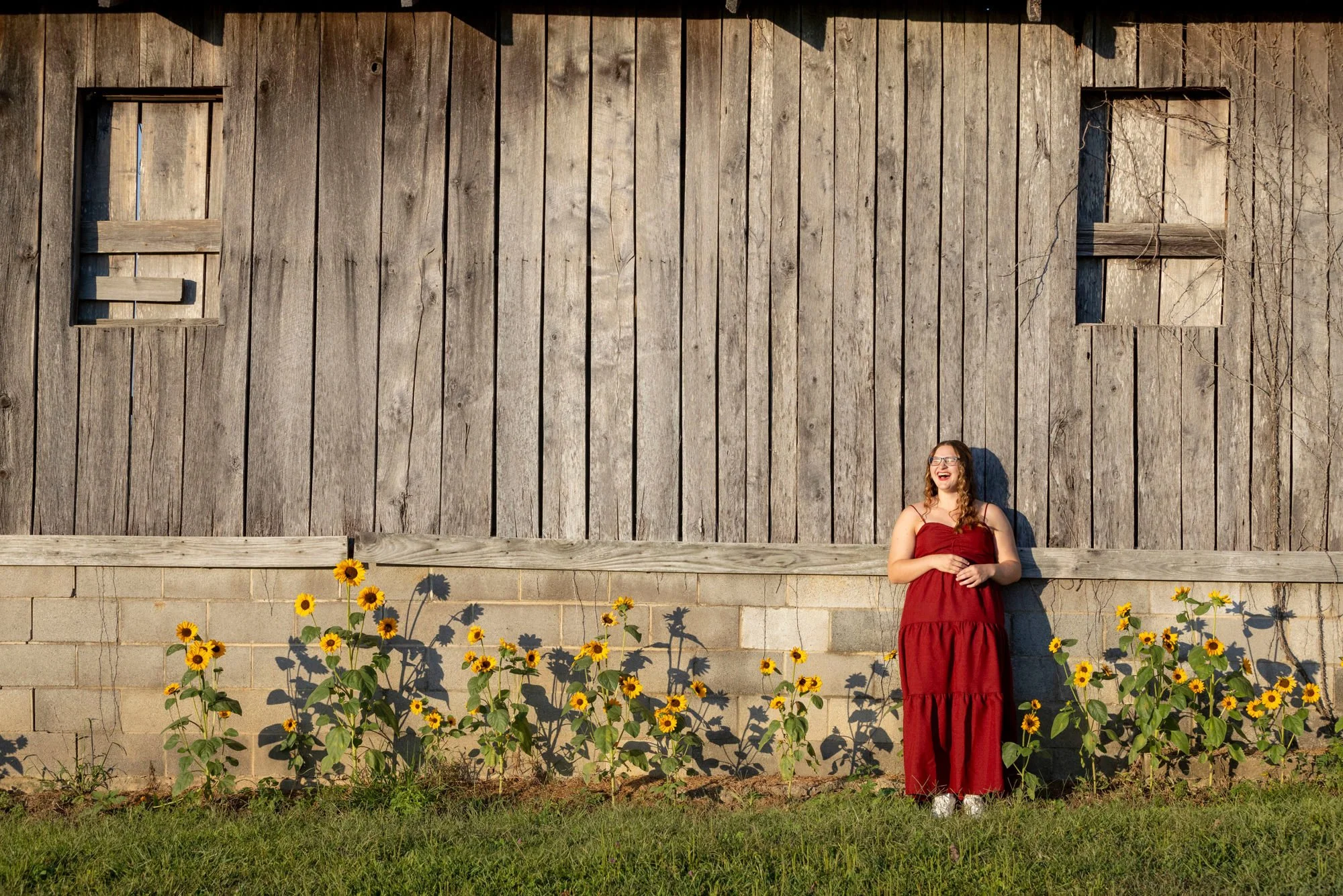 A young woman with glasses and long, curly hair wearing a long red dress, smiling and standing in front of a large wooden barn wall with two boarded-up windows. She is standing on green grass with yellow sunflowers growing in front of her, and the su