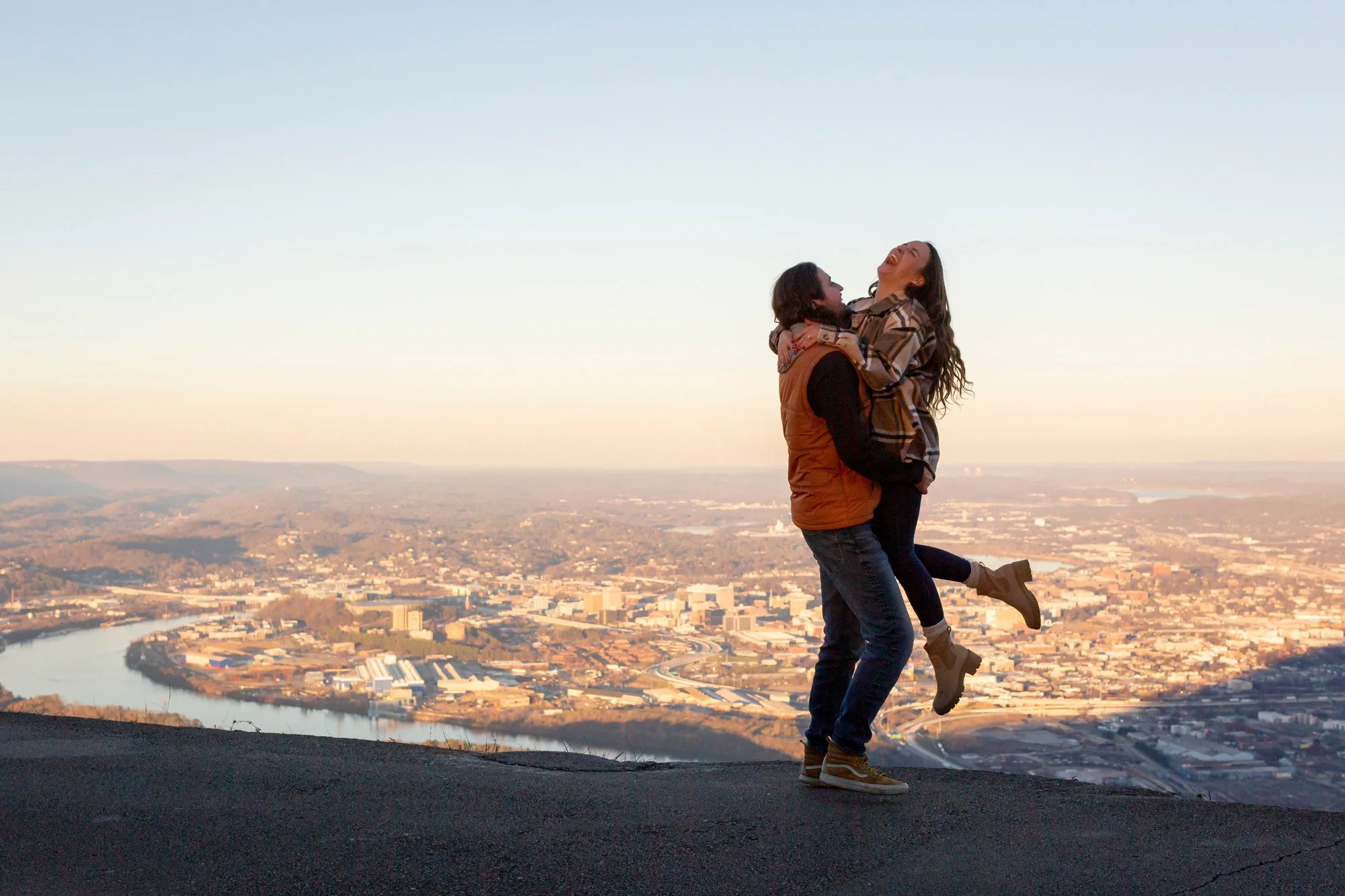 A happy couple in casual clothing, with the man lifting the woman in celebration, standing on a hill overlooking a cityscape beneath a clear sky at sunset.