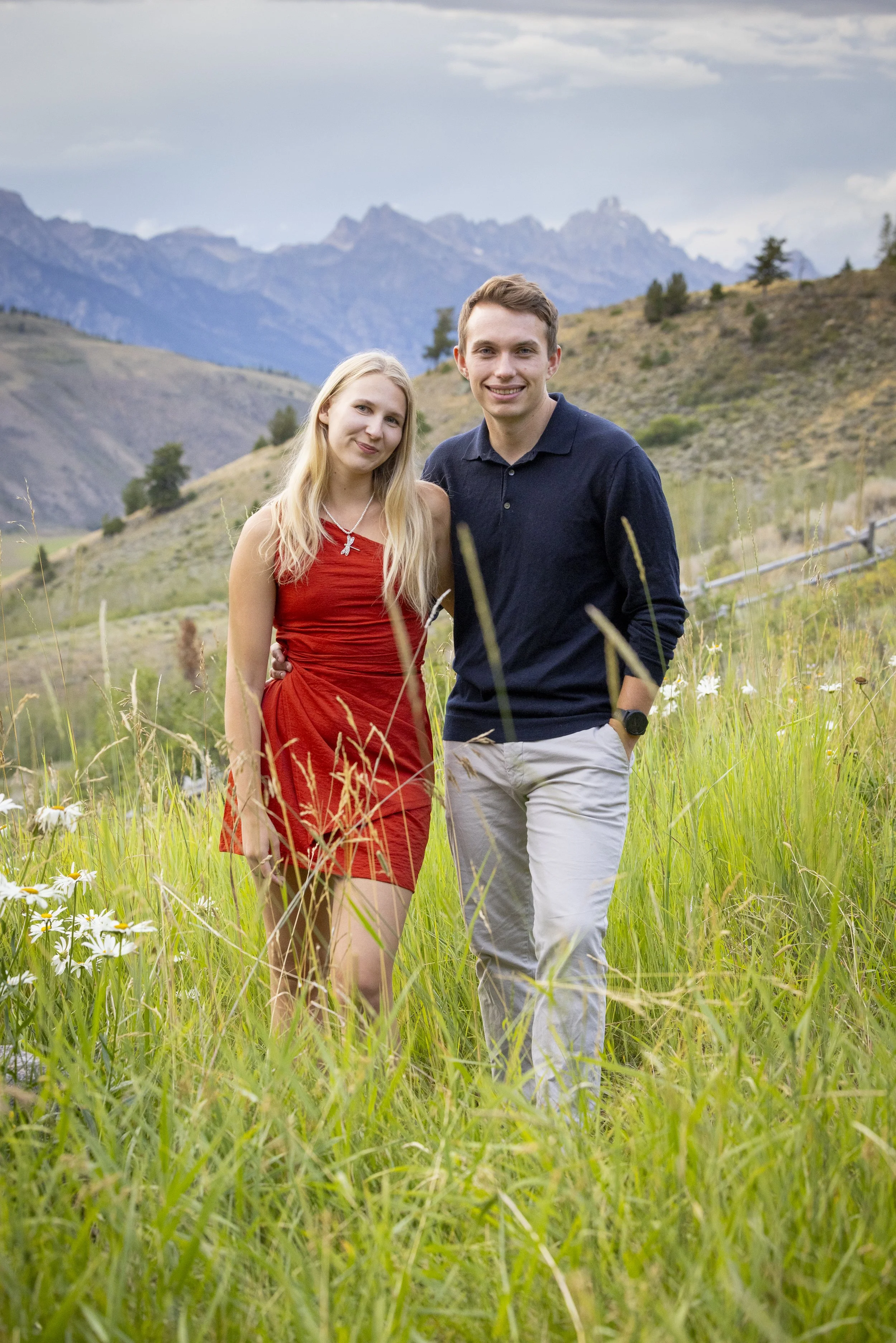 A young man and woman standing together in a grassy field with wildflowers, mountains in the background, under cloudy skies, smiling at the camera.