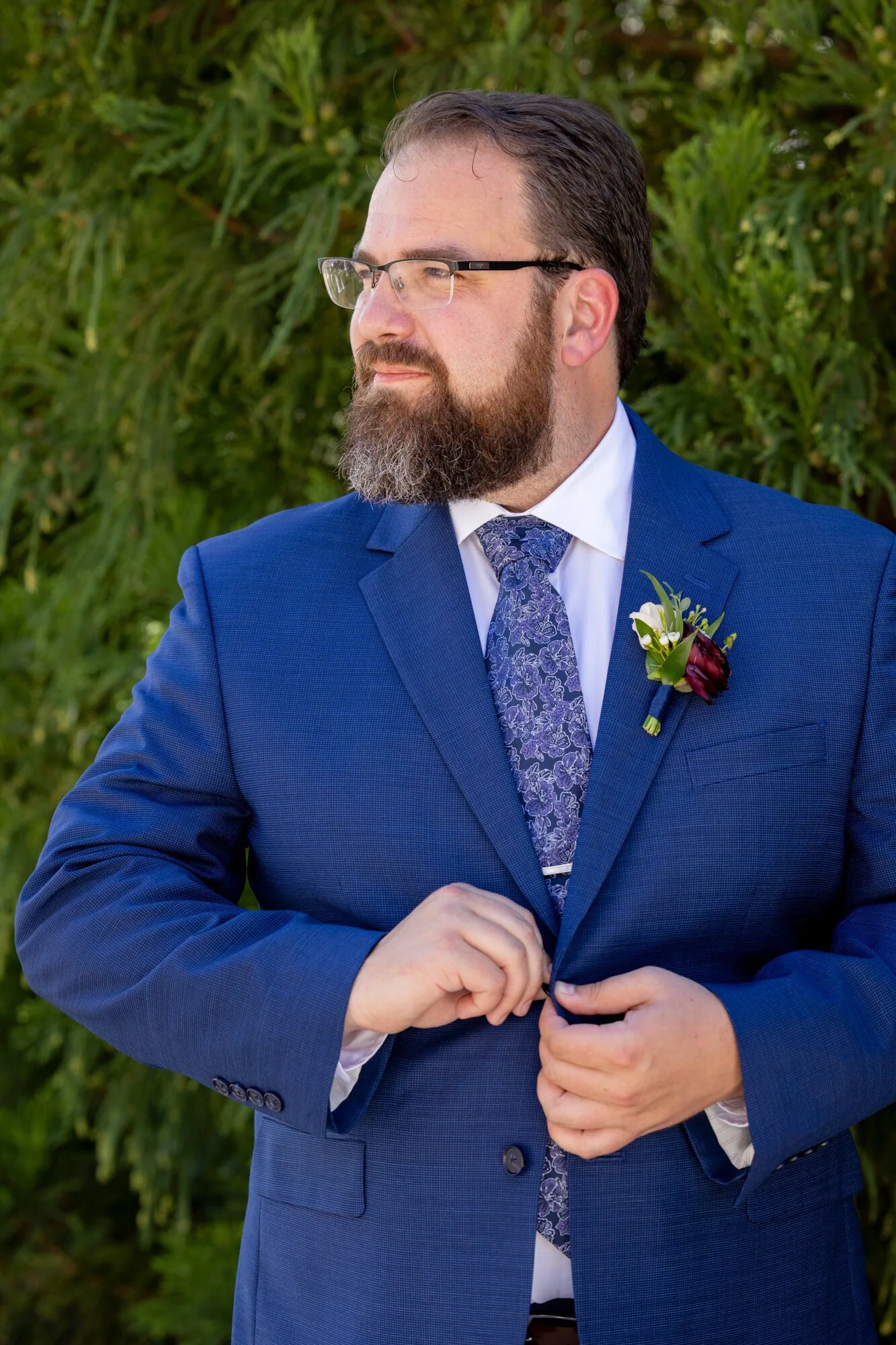A man in a blue suit with a floral tie and boutonniere stands outdoors in front of green foliage, with glasses and a beard, looking to his left.
