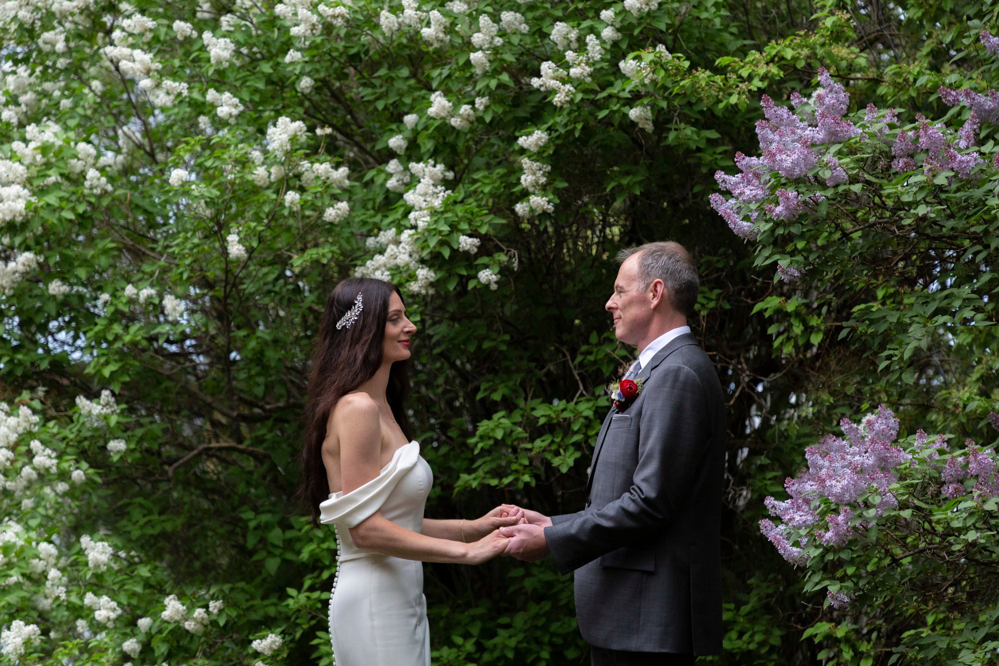 A bride and groom holding hands during their wedding ceremony outdoors, surrounded by lush green foliage and blooming purple and white flowers.