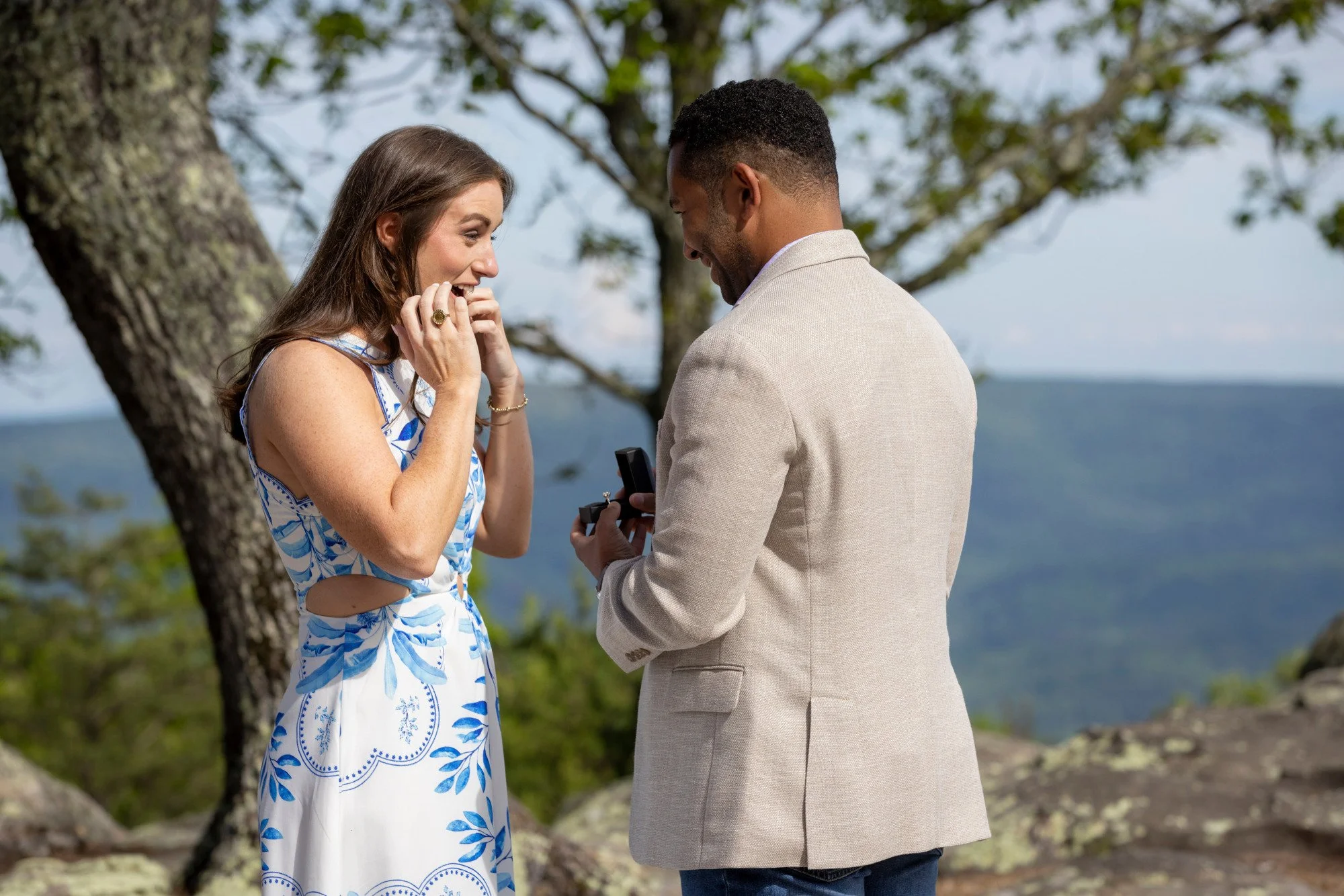 A man is proposing marriage to a woman outdoors by a lake or ocean, with trees in the background. The woman is surprised and emotional, holding her hands near her face, while the man is smiling and holding an open jewelry box.