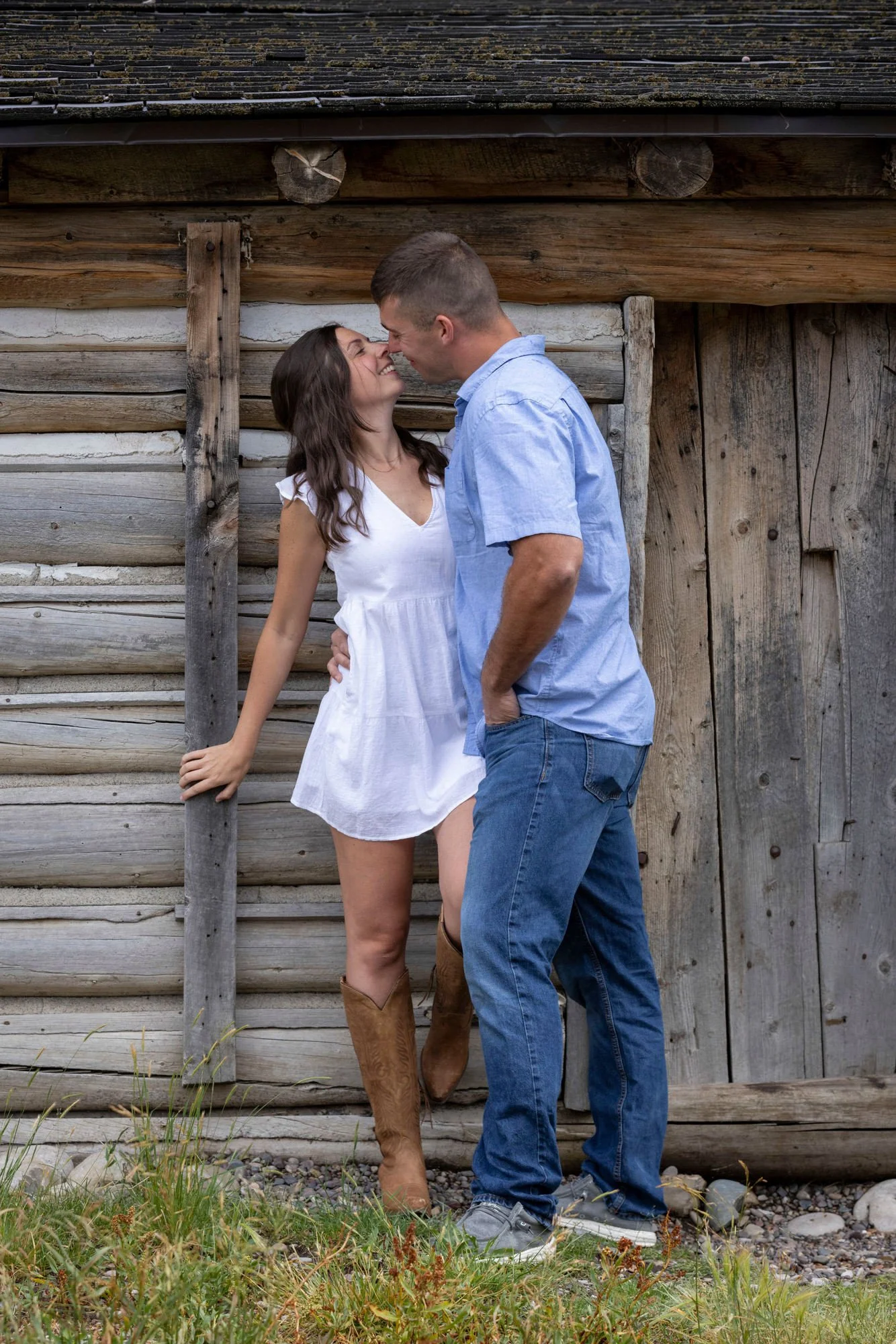 A couple standing close to each other in front of a wooden barn wall, about to kiss, with the woman wearing a white dress and cowboy boots, and the man in a light blue shirt and jeans.