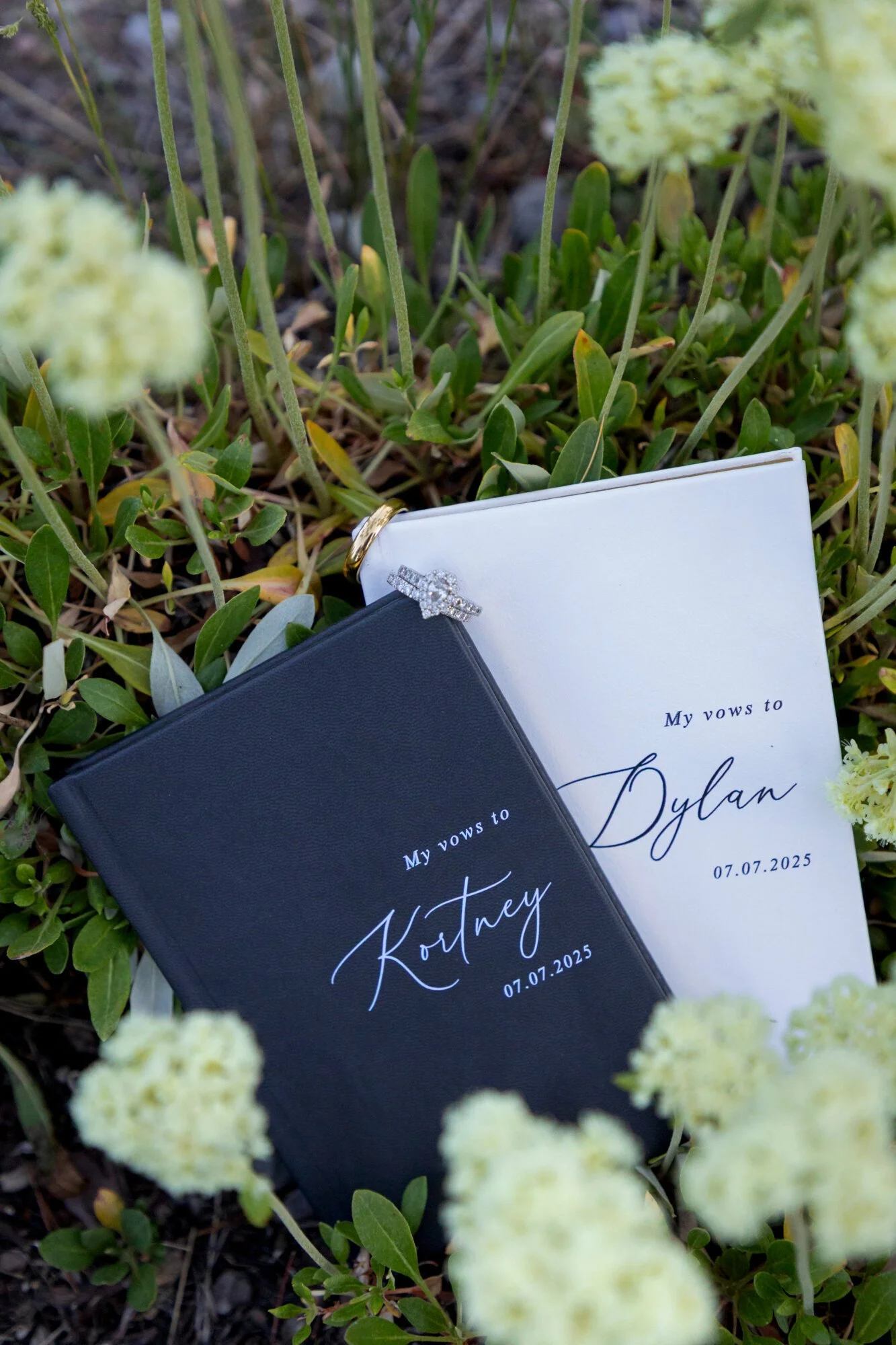 A black and a white booklet with the words 'My vows to' followed by handwritten names, and a date '07.01.2025,' placed on green grass among white flowers.