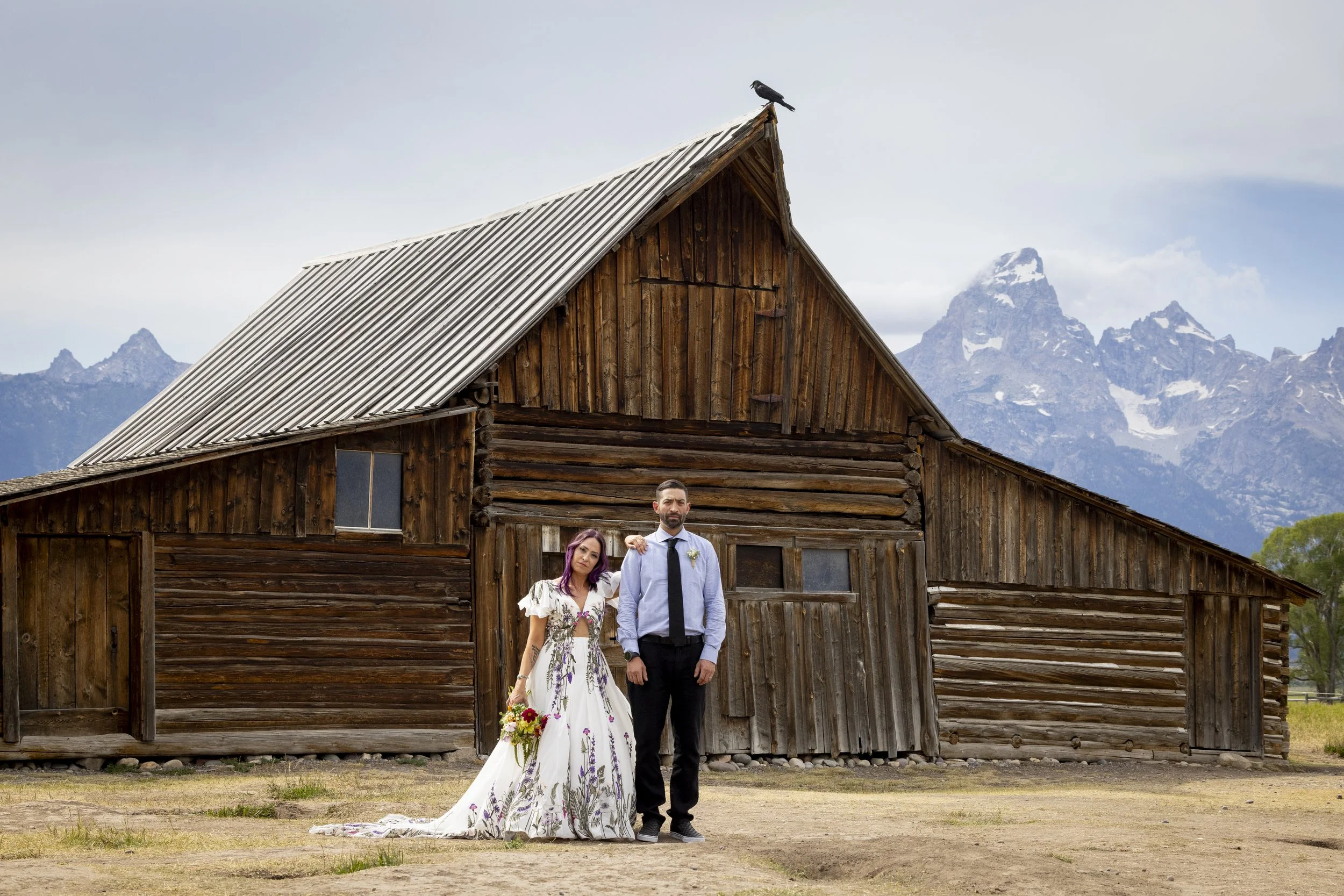 A bride and groom standing in front of a rustic wooden barn with mountain peaks in the background. The bride is wearing a white dress with floral patterns and holding a bouquet, while the groom is wearing a light blue shirt, black tie, and dark trous