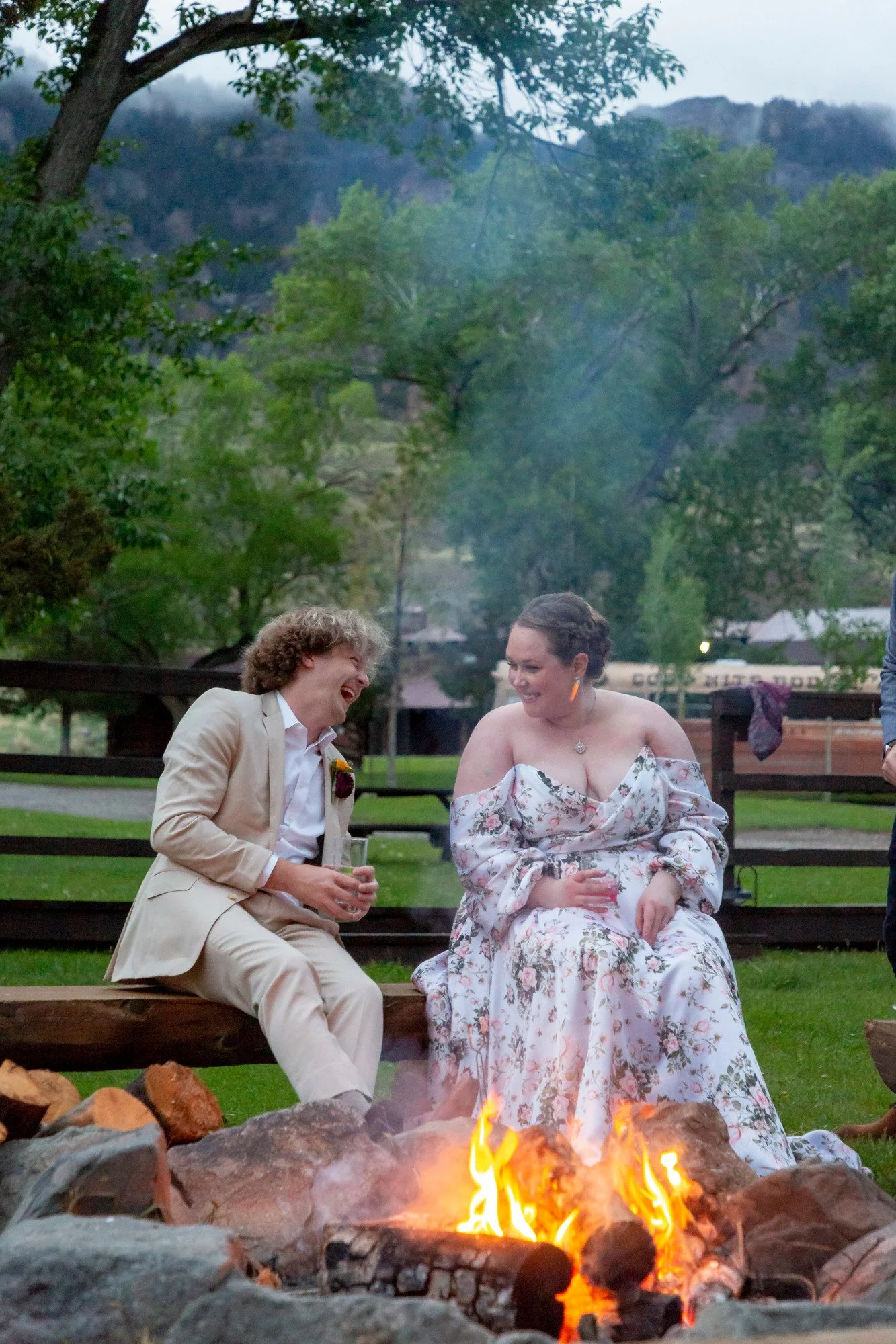 A couple sitting by a campfire outdoors, laughing and smiling, with trees and hills in the background, during evening or dusk.