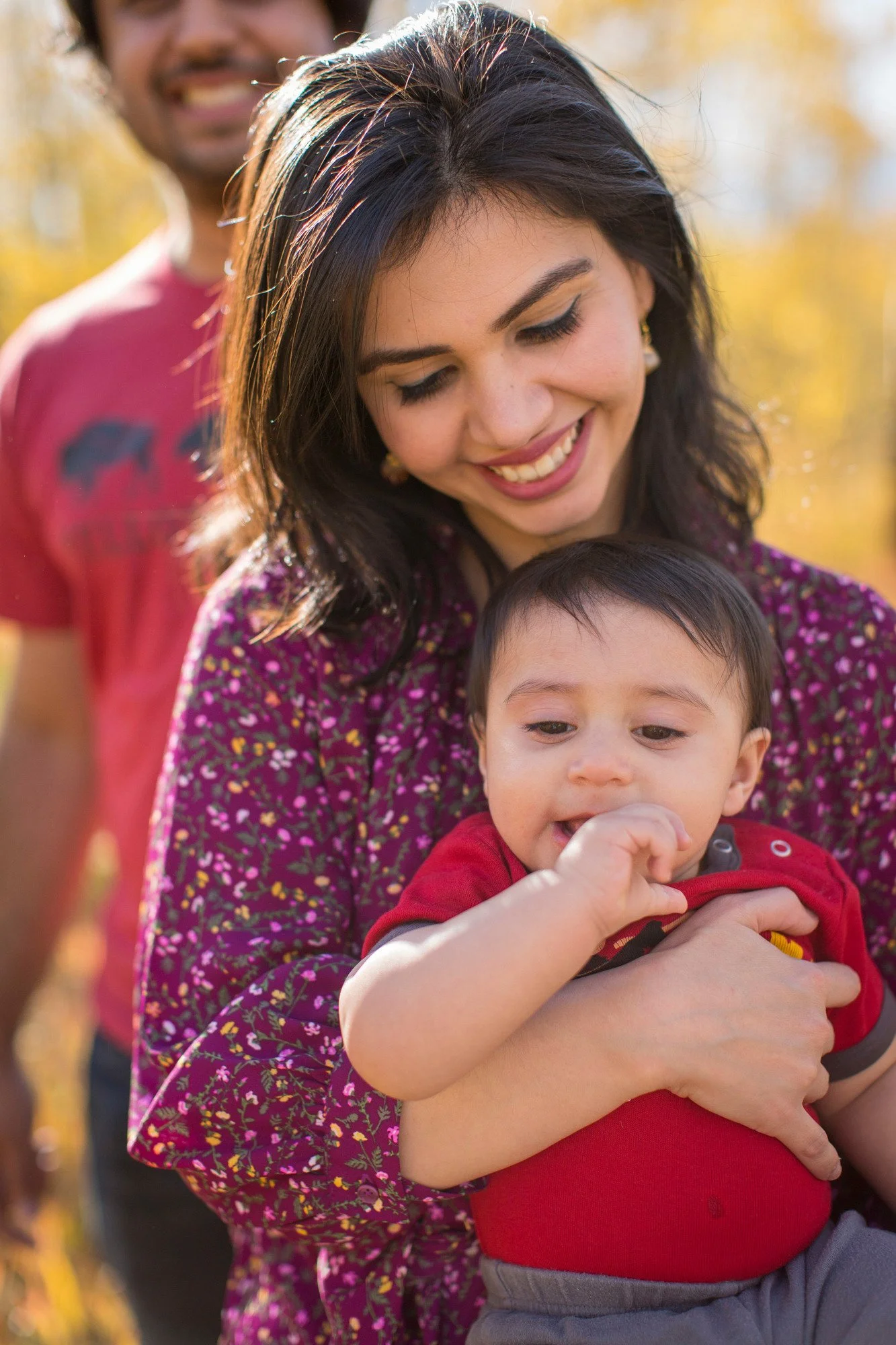 A woman is smiling while holding a young child who is wearing a red shirt and appears to be eating or chewing on their hand, with a blurry adult man smiling in the background outdoors with trees displaying fall colors.