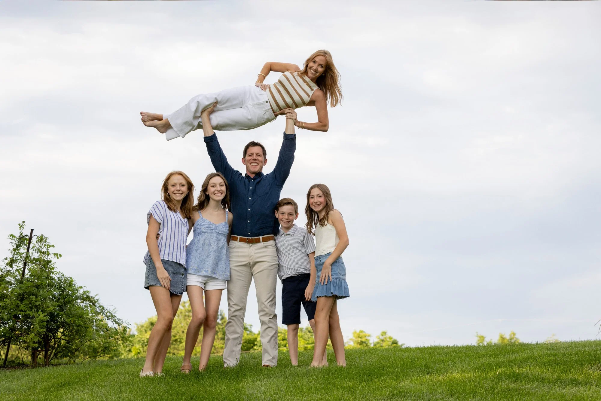A man in a navy blue shirt and beige pants holding a woman in a striped top and white pants above his head, surrounded by four young girls on grass with trees in the background and a cloudy sky.