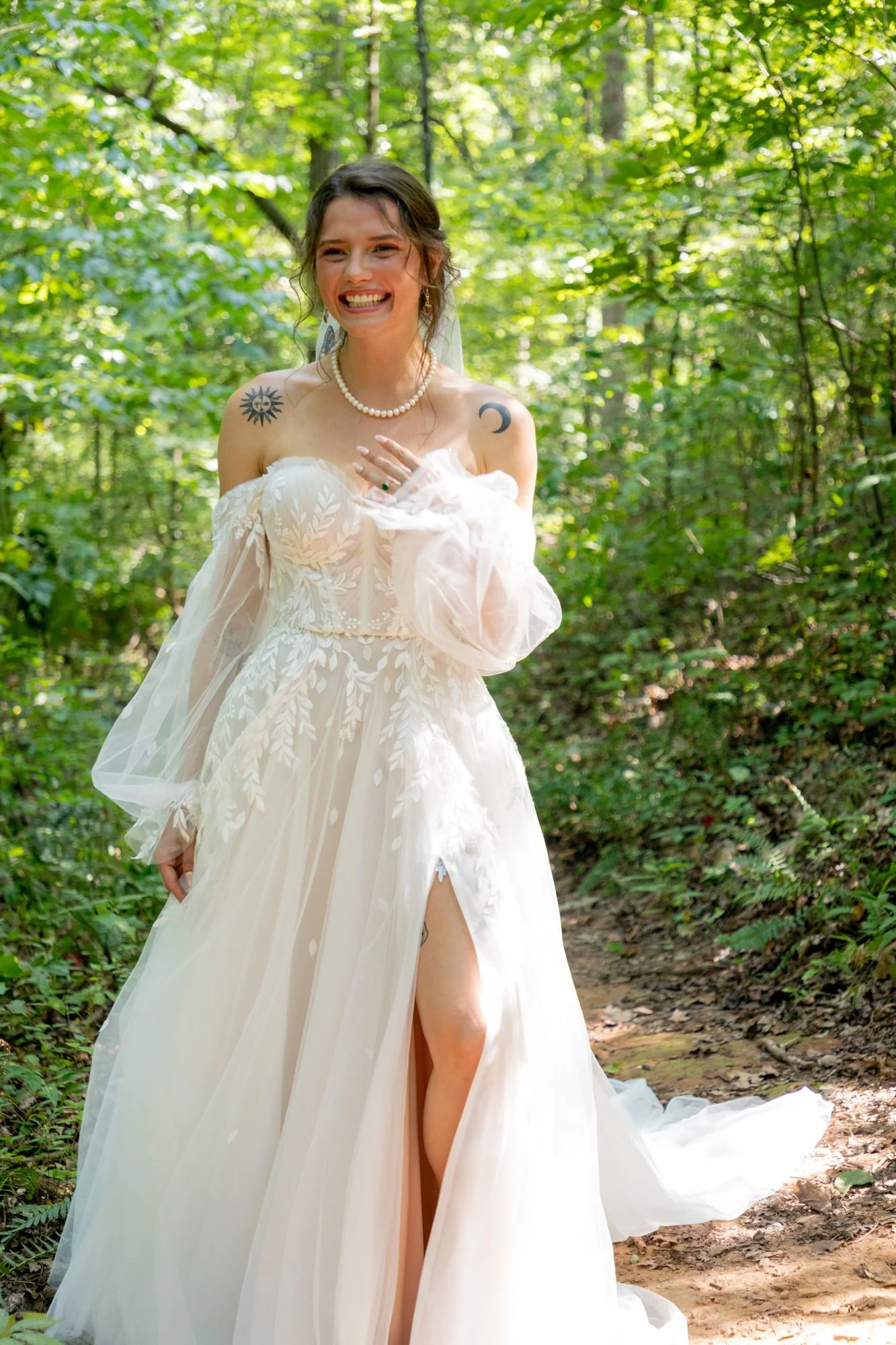 A smiling woman in a white wedding gown standing in a lush green forest, with tattoos visible on her shoulders and a pearl necklace.