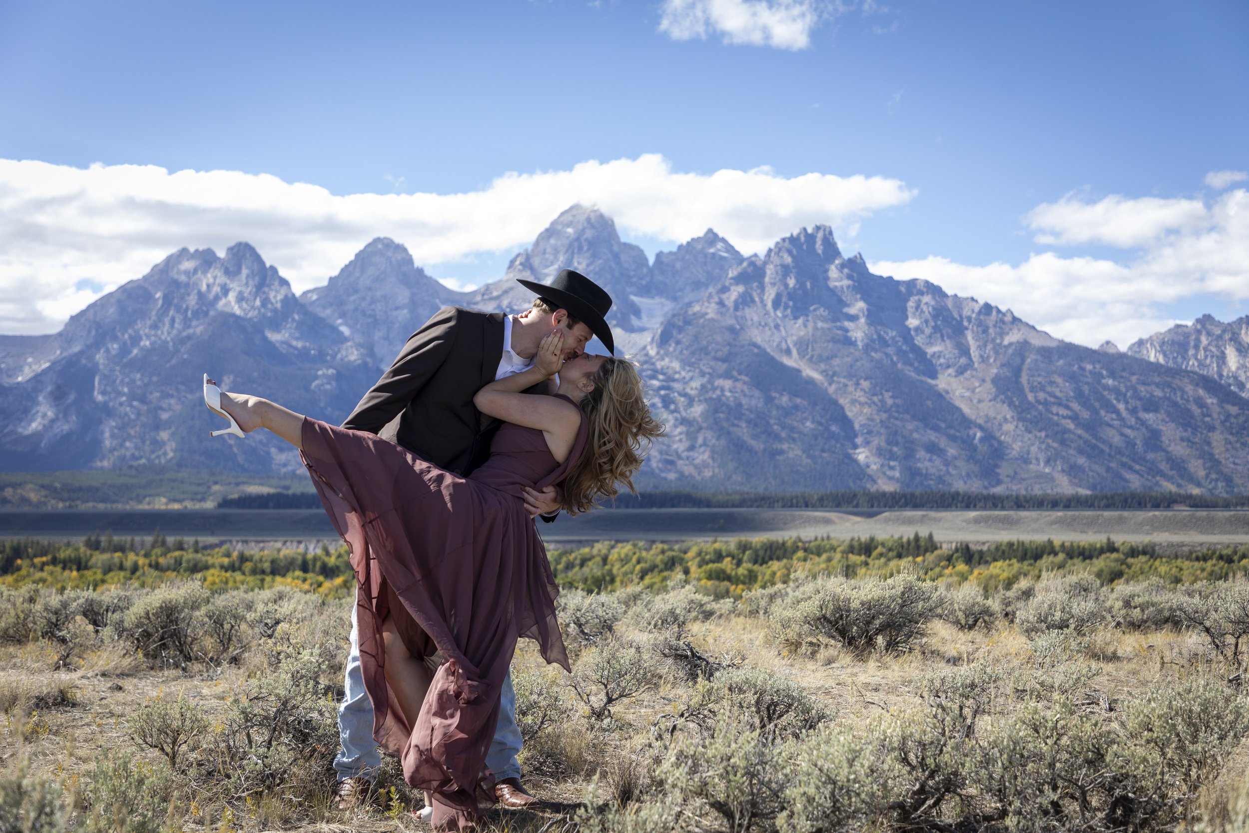 A couple dressed formally in a romantic pose with the man lifting the woman in a desert landscape with mountains in the background.