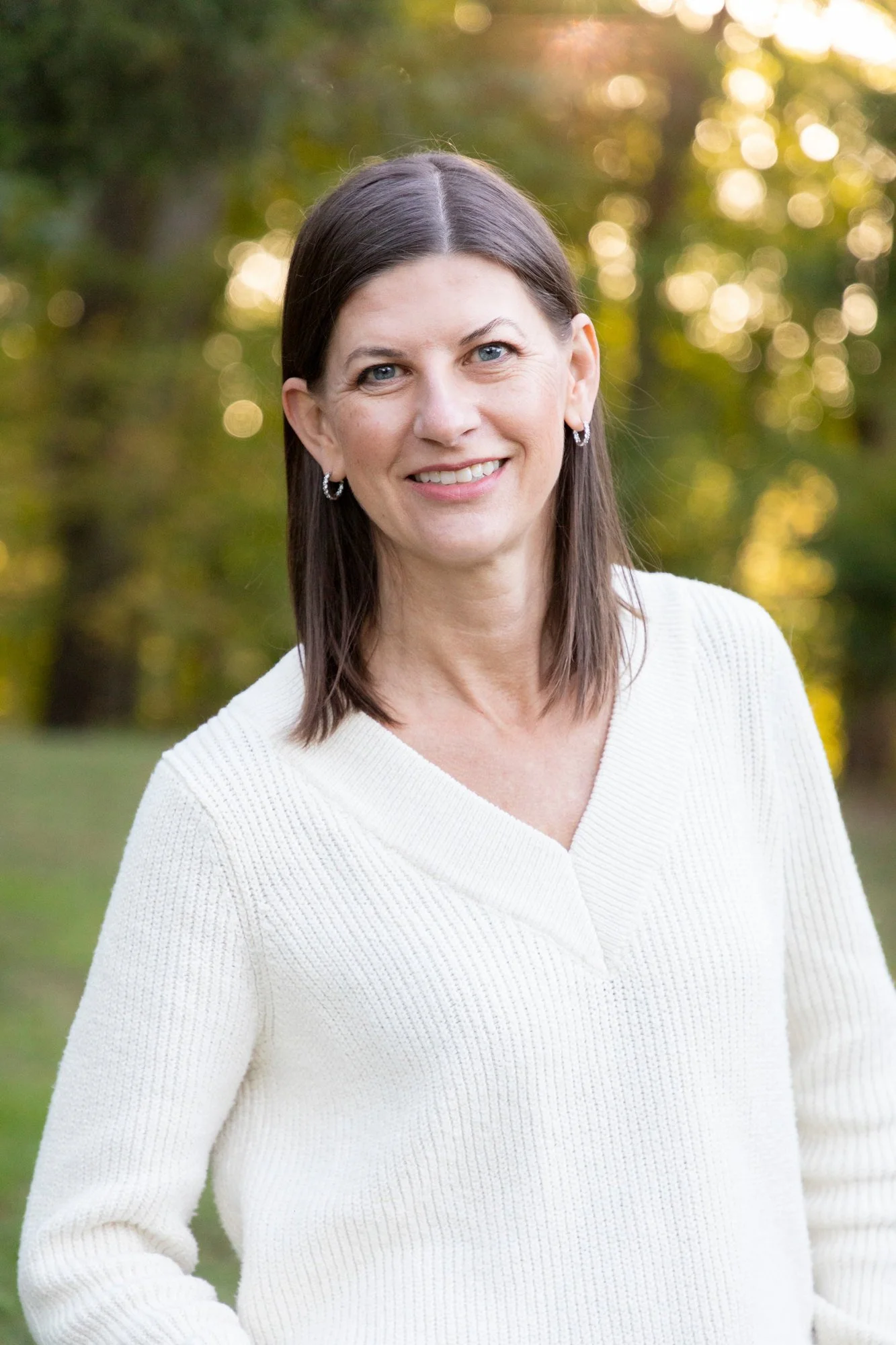 A woman with dark brown hair and blue eyes smiling outdoors, wearing a white knitted sweater and small hoop earrings, with blurred trees and sunlight in the background.