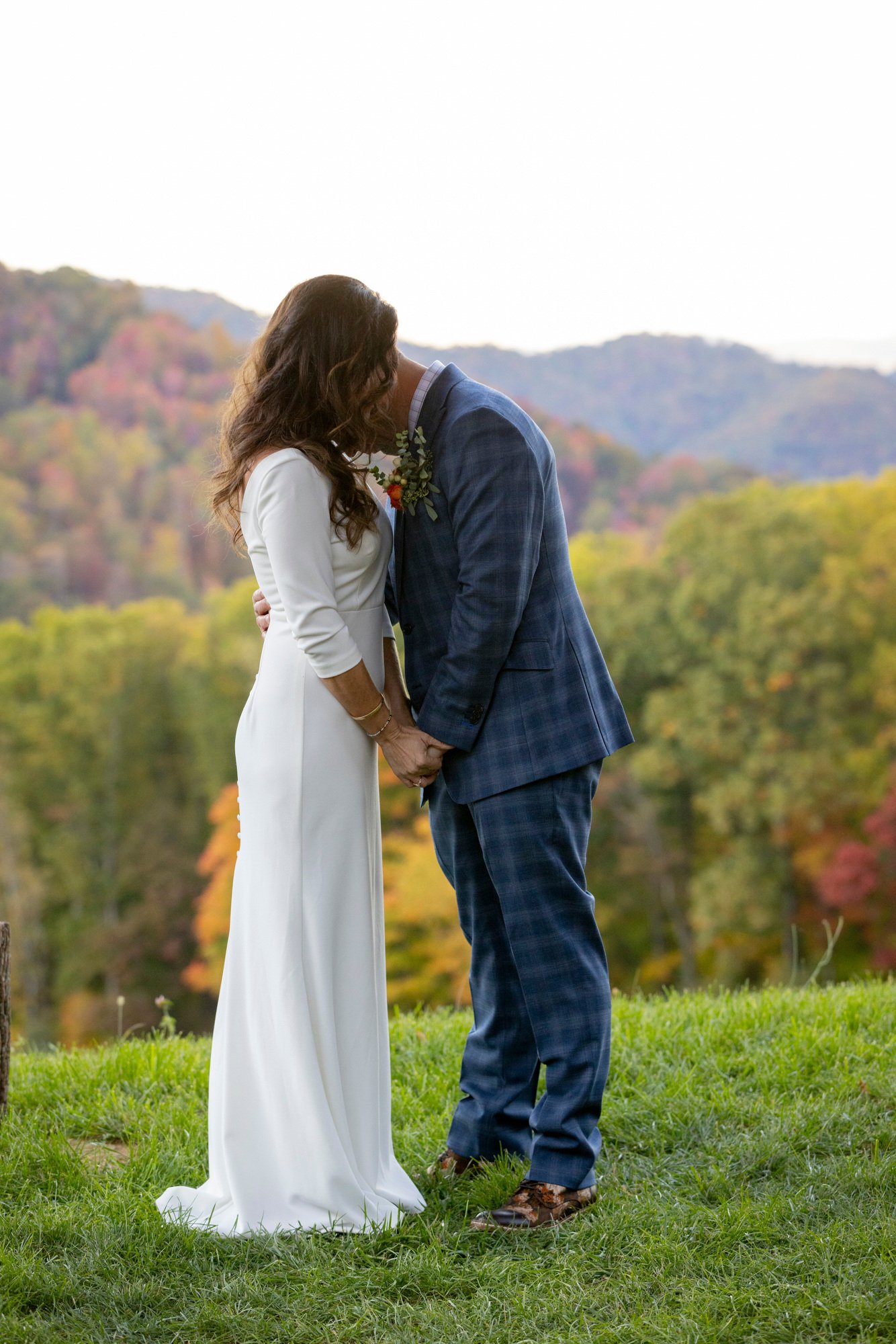 A couple dressed in wedding attire sharing a kiss outdoors with forested hills in the background.