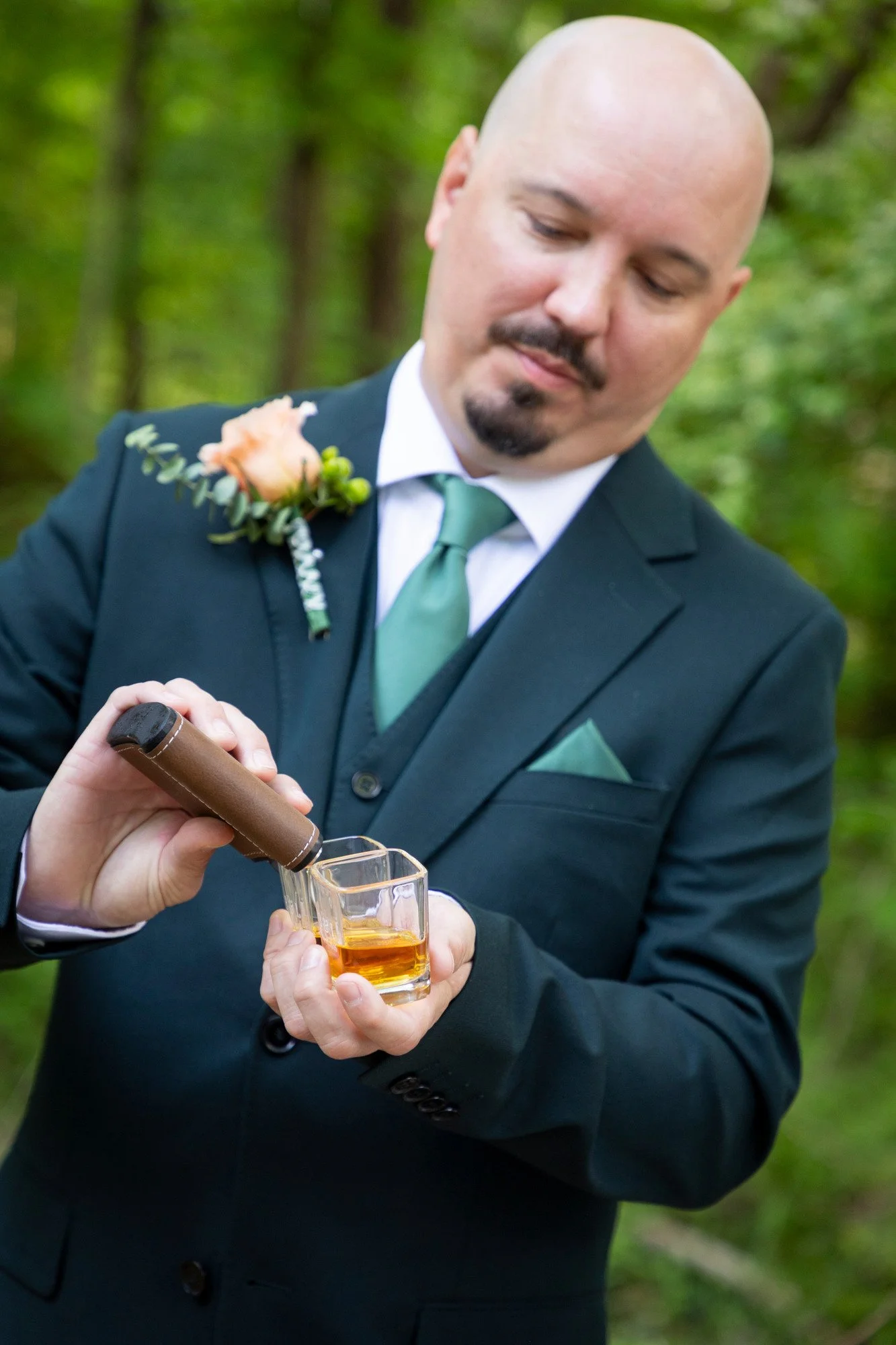 A man in a black suit and green tie pouring a drink from a bottle into a glass held by another person, outdoors with green trees in the background.