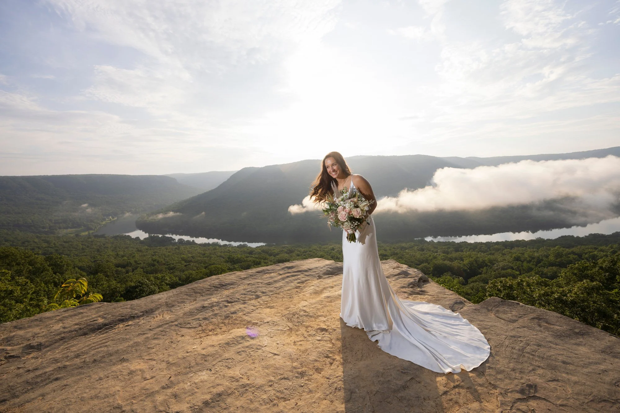 A woman in a white wedding dress holding a bouquet of flowers, standing on a rocky cliff overlooking a green valley with a river and mountains under a partly cloudy sky.