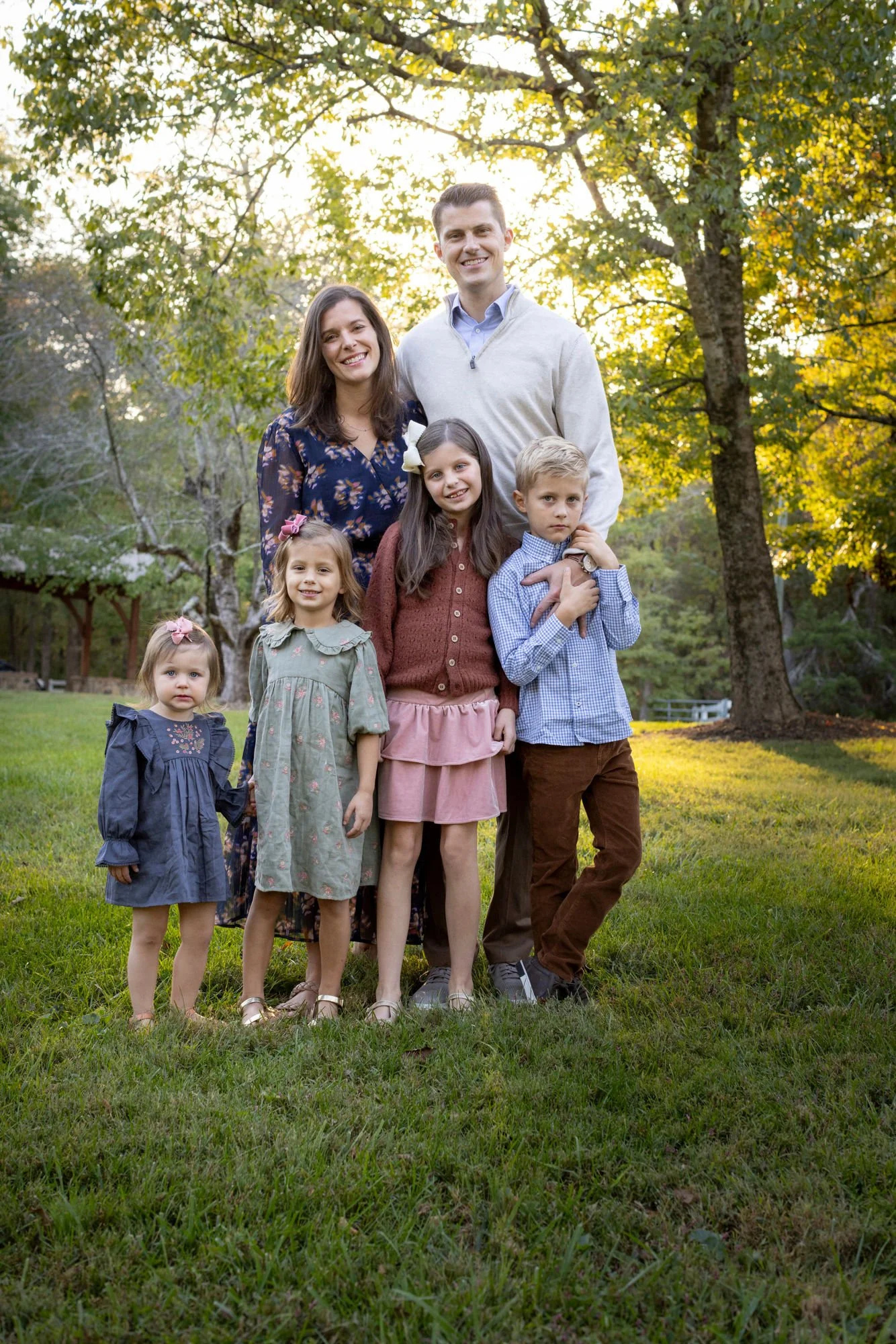 A family of seven standing outdoors in a park during golden hour. Two adults, three girls, and two boys, all smiling and dressed casually. Behind them are large trees with green and yellow leaves.