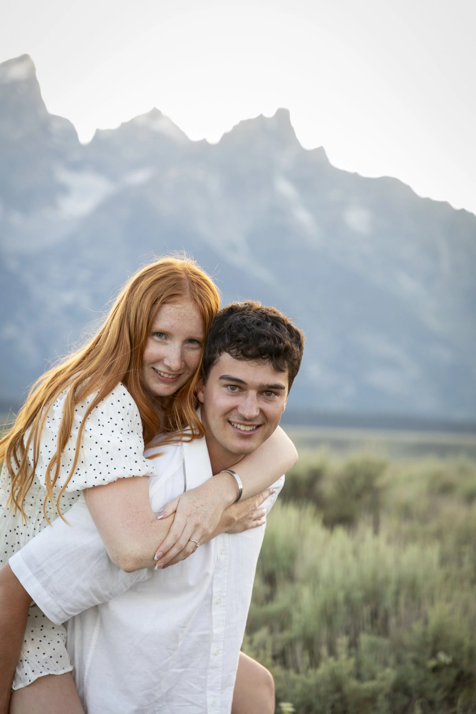 A young couple, a woman with long red hair and a man with short dark hair, smiling and hugging outdoors with mountains in the background.