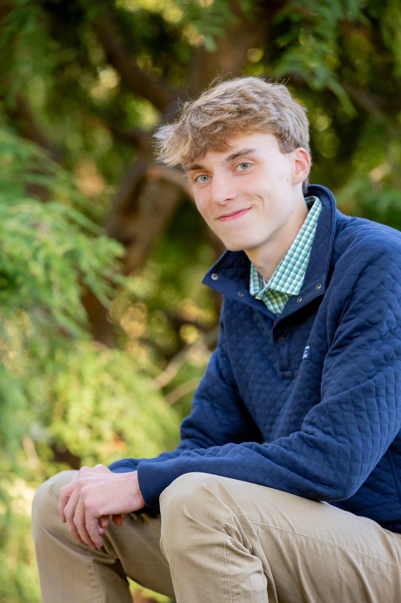 Young man with light brown hair and blue eyes sitting outdoors on a tree branch, smiling, wearing a blue quilted jacket and beige pants, with green foliage in the background.