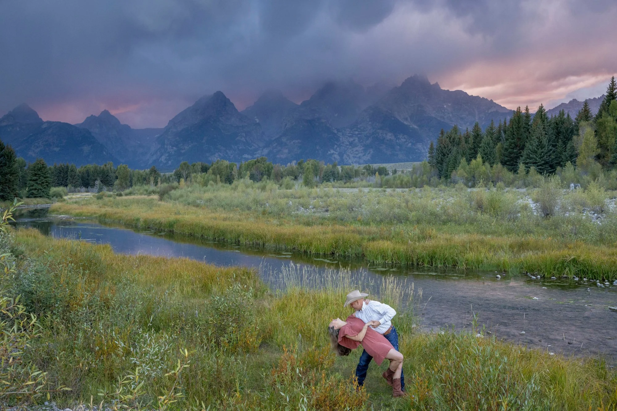 A couple dancing by a river in a lush, green landscape with tall trees, mountains in the background, and cloudy skies at sunset.