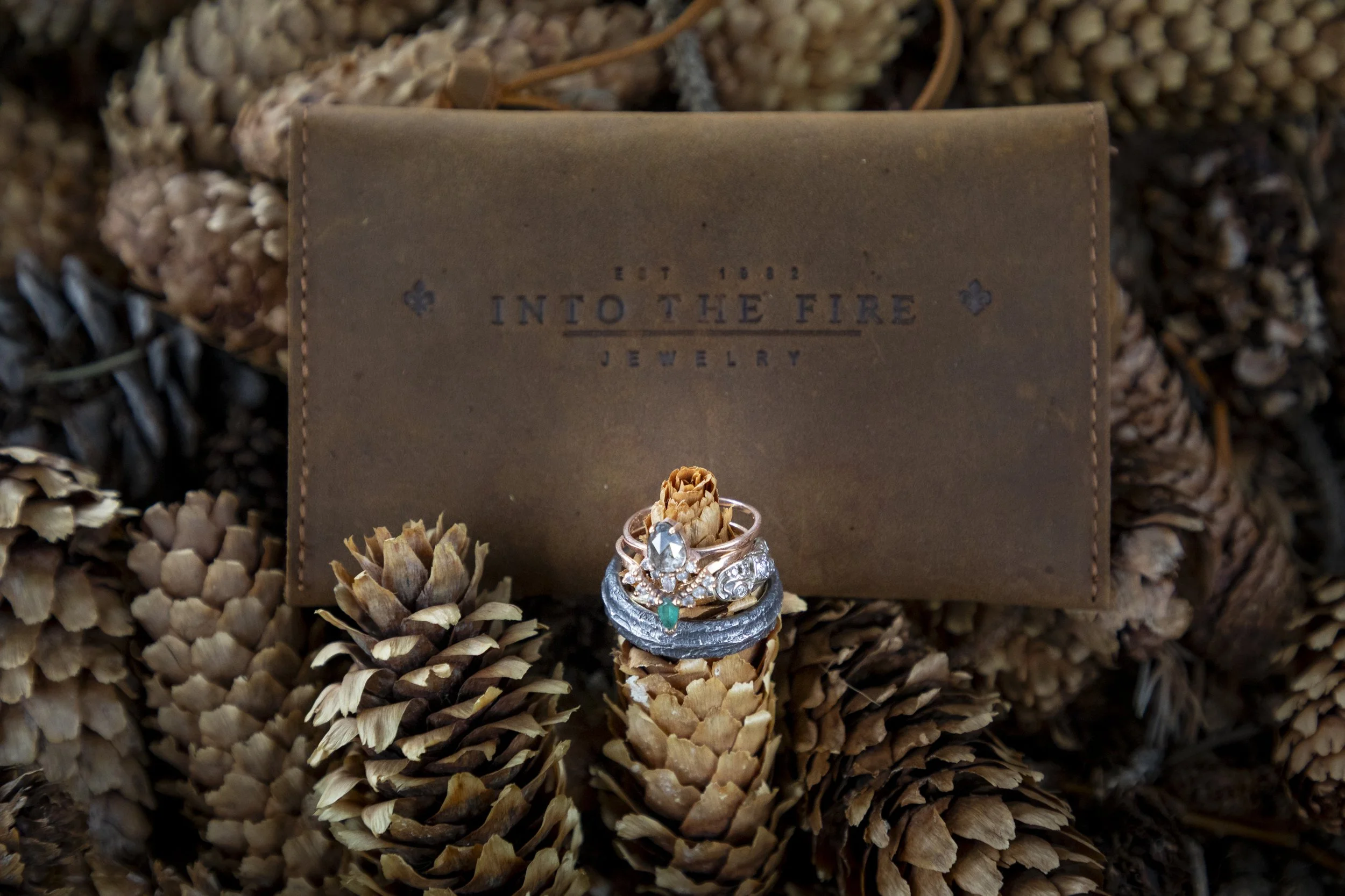 A jewelry box on a bed of pinecones contains a set of rings and a small decor piece. The jewelry box has the words 'Into the Fire Jewelry' printed on it.