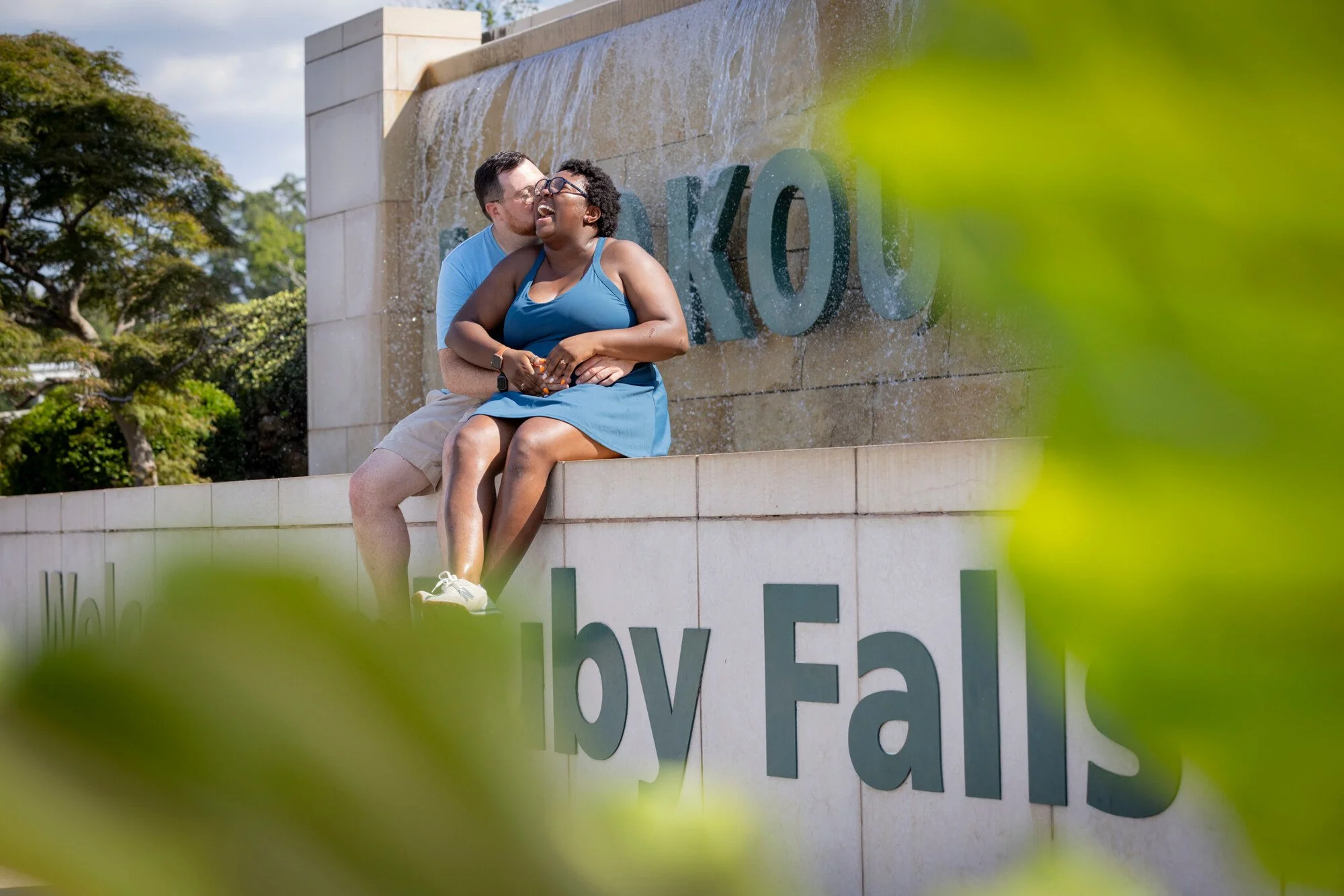 A person kissing another person on the cheek in front of a water fountain, with a large sign and trees in the background, partly obscured by green leaves in the foreground.