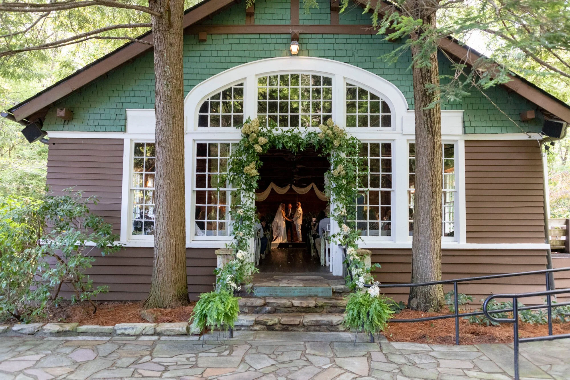 A green and brown house with large windows and an arch entrance decorated with white flowers and greenery, surrounded by trees during daytime.