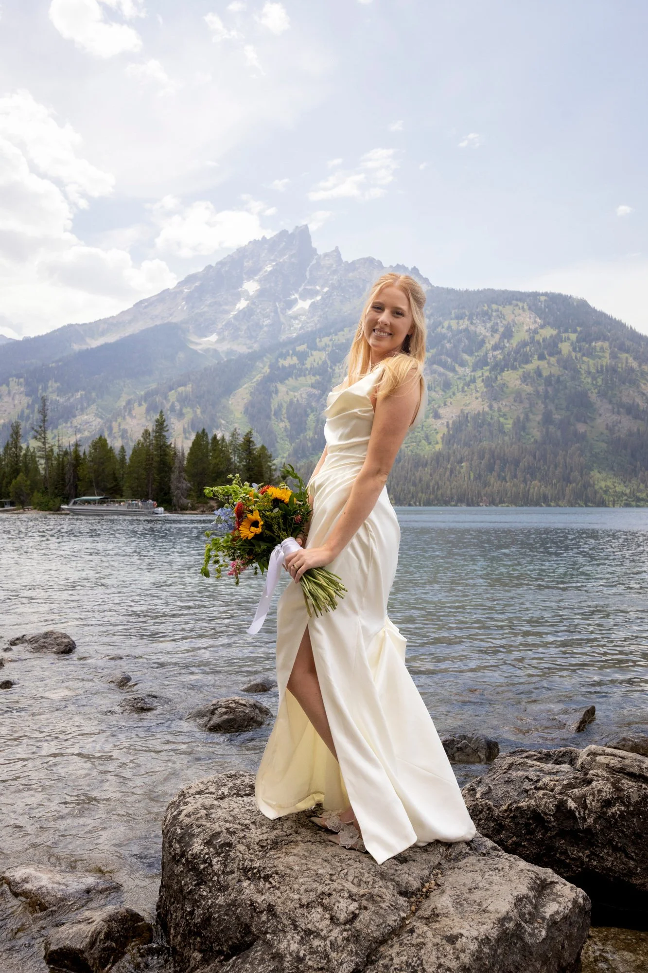 A woman in a white wedding dress holding a colorful bouquet standing on rocks by a lake with mountains and trees in the background.