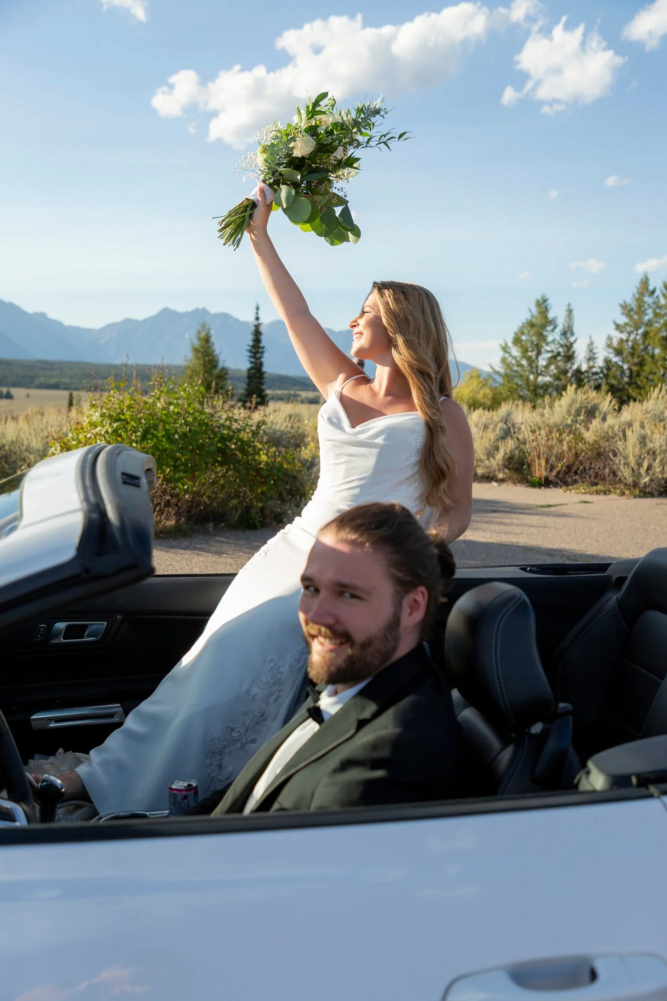 A bride in a white wedding dress and a groom in a tuxedo sitting in a convertible car, with the bride holding a bouquet of flowers up in the air and both smiling. The background features mountains, trees, and a blue sky with some clouds.