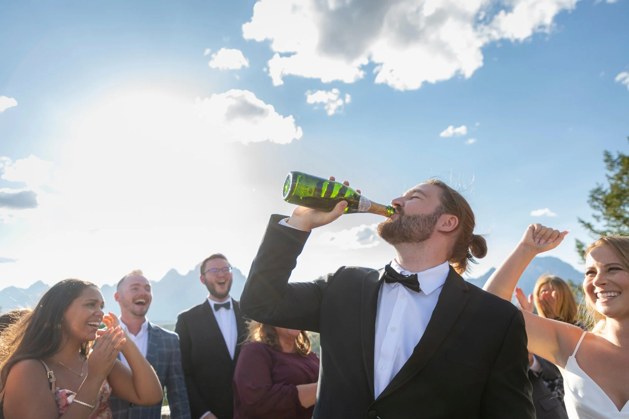 A man in a tuxedo and bow tie drinks champagne from a bottle at an outdoor celebration, with smiling people around him and mountainous scenery in the background.