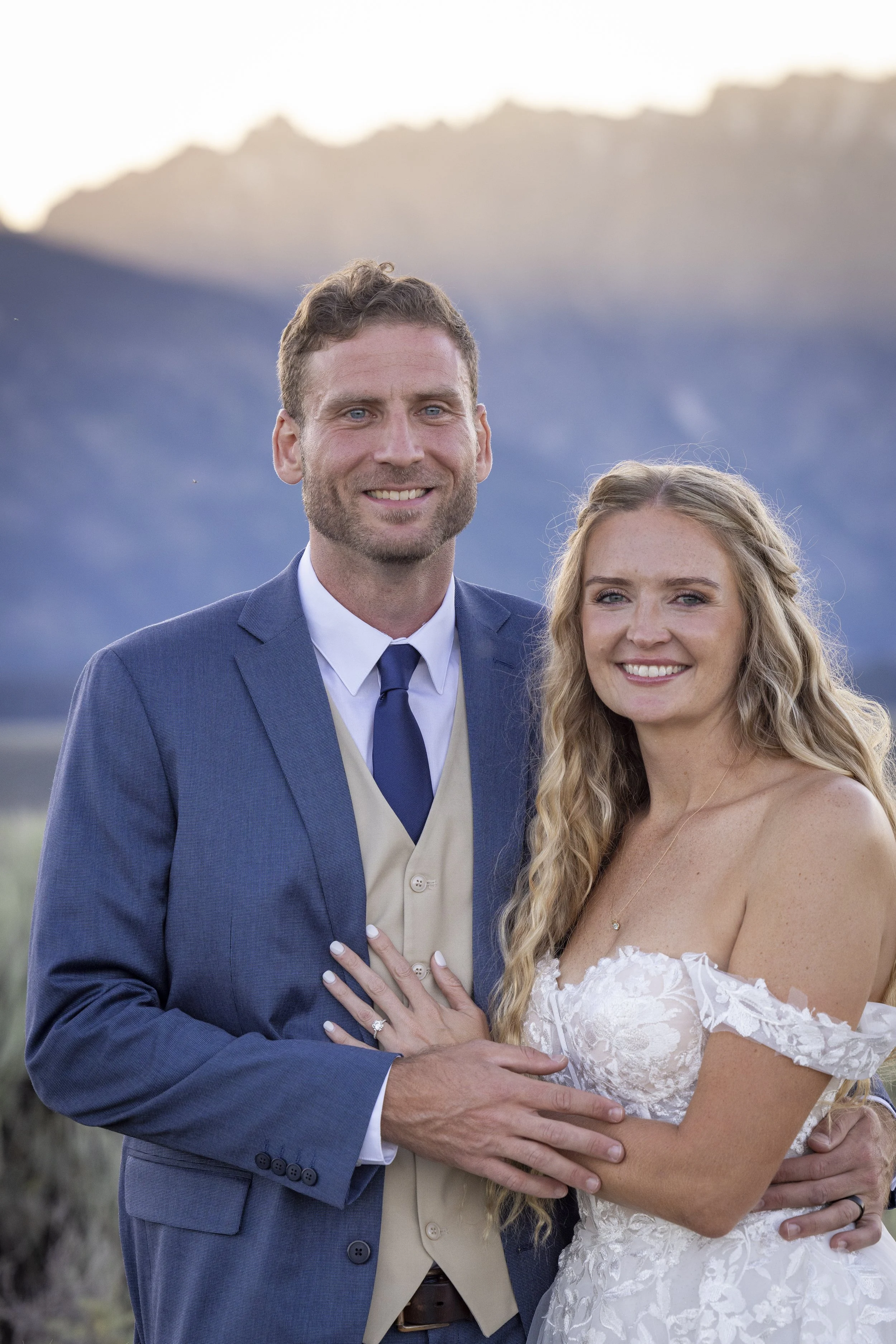 A happy couple dressed in wedding attire, standing outdoors with mountains in the background during sunset.
