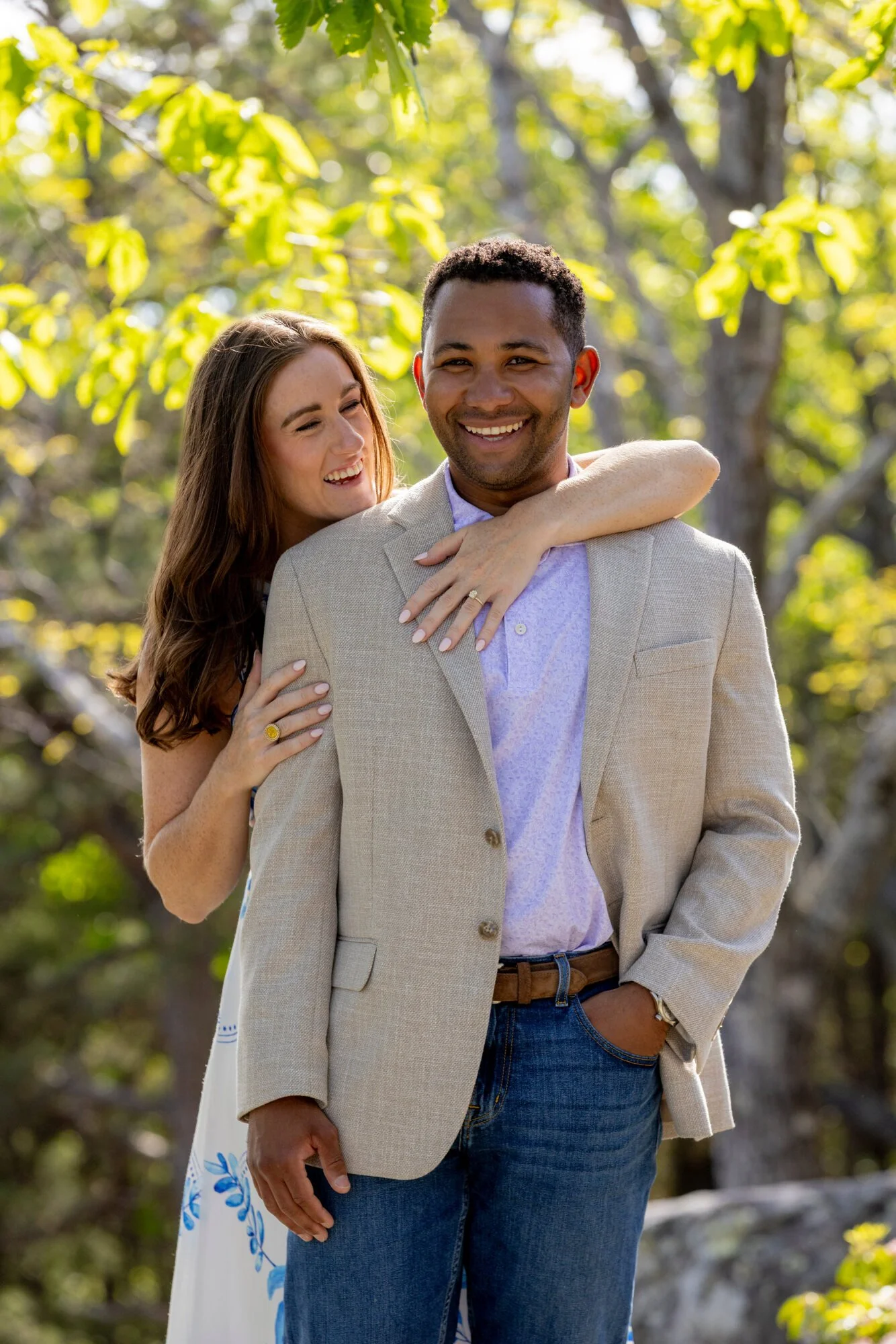 A happy couple outdoors in a park, with the woman embracing the man from behind. The man is wearing a beige blazer and jeans, and the woman has long brown hair and is smiling, wearing a white dress with blue floral patterns. The background features g