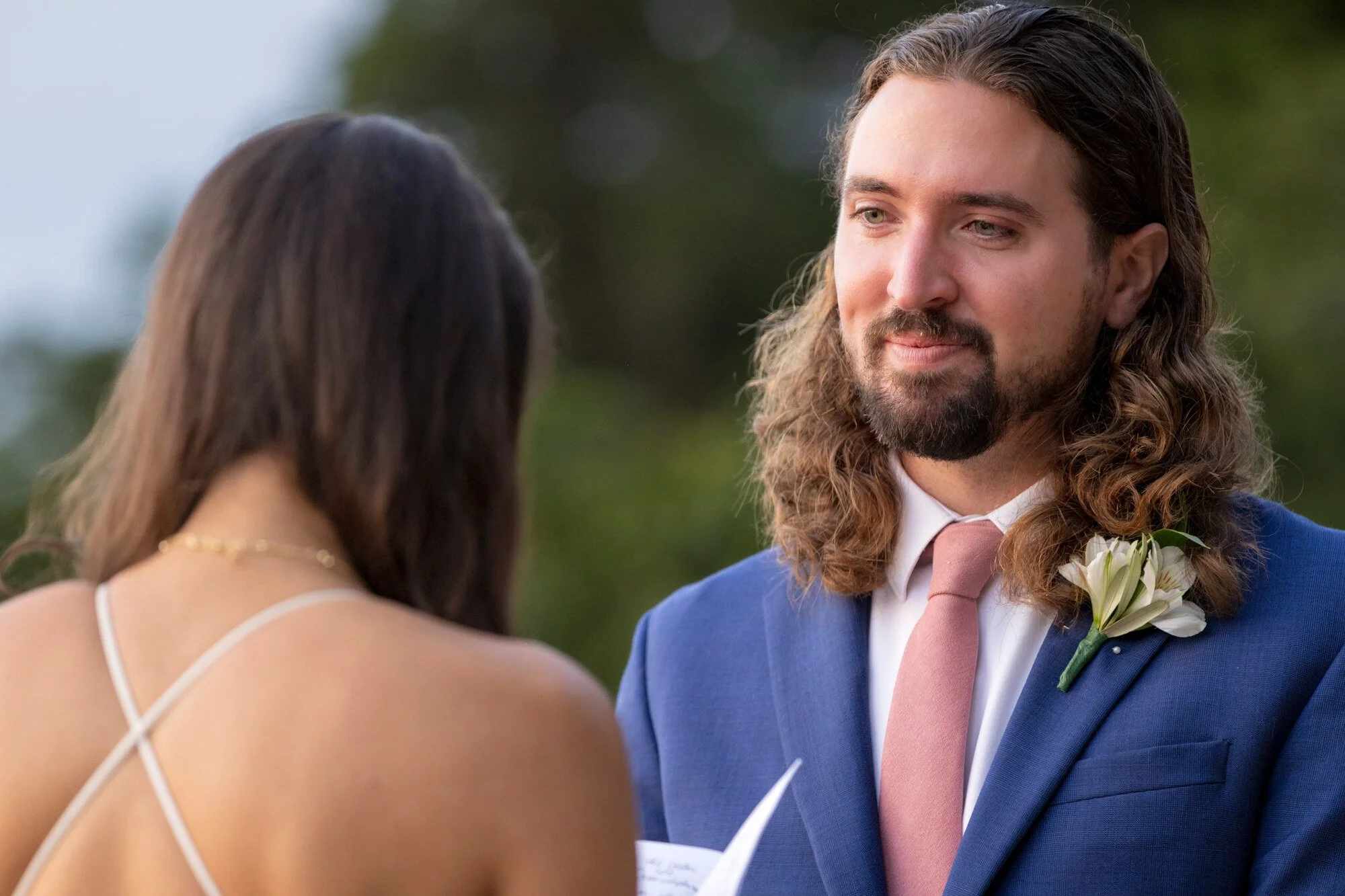 A man with long, wavy hair and a beard wearing a blue suit and pink tie is looking at a woman. The woman, who has long brown hair and is wearing a white dress with thin straps, is facing him. The scene appears to be outdoors with green foliage in the