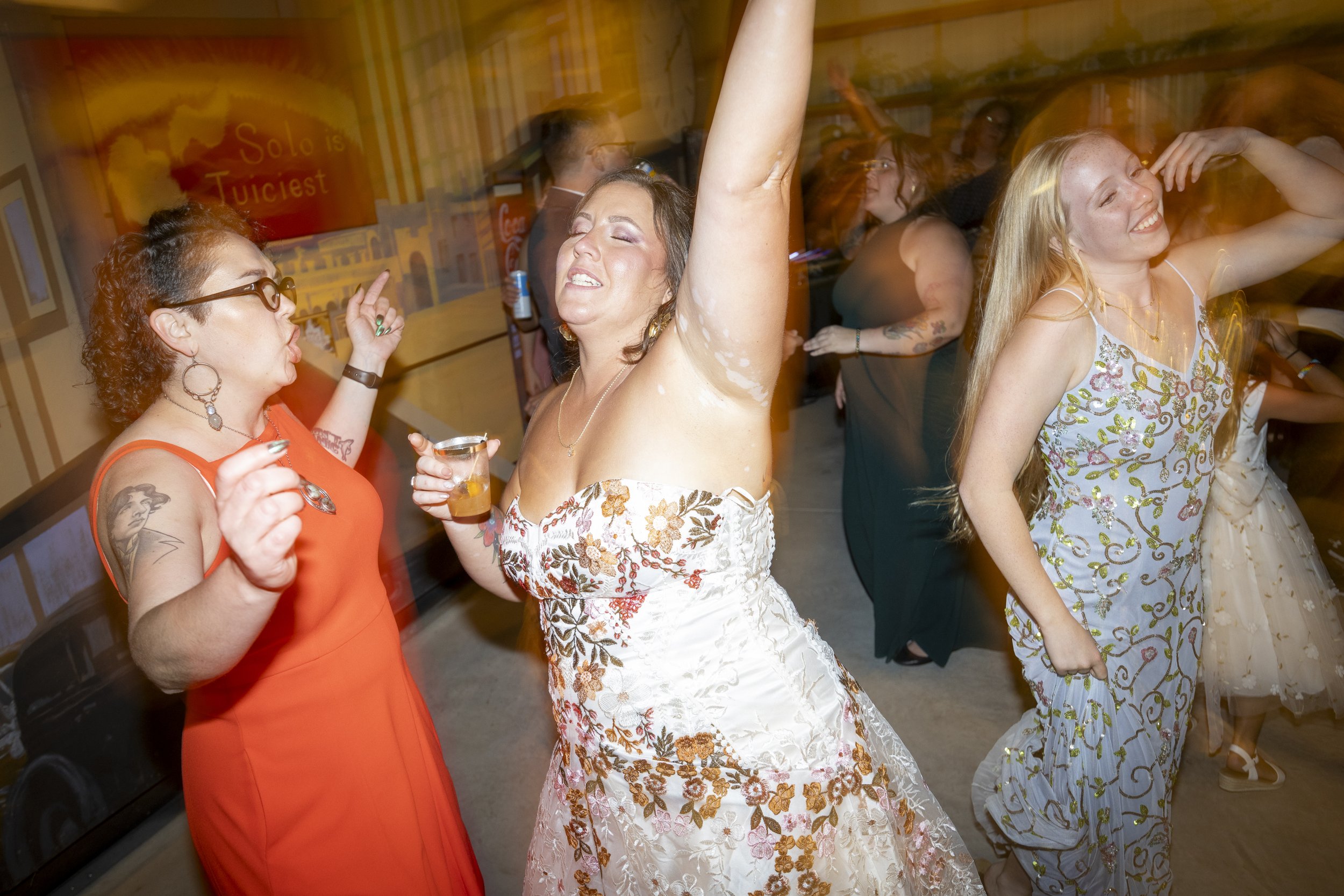 People dancing and enjoying themselves at a party or celebration, with a woman in a white floral dress and two women in colorful dresses, one orange and one blue with floral patterns.