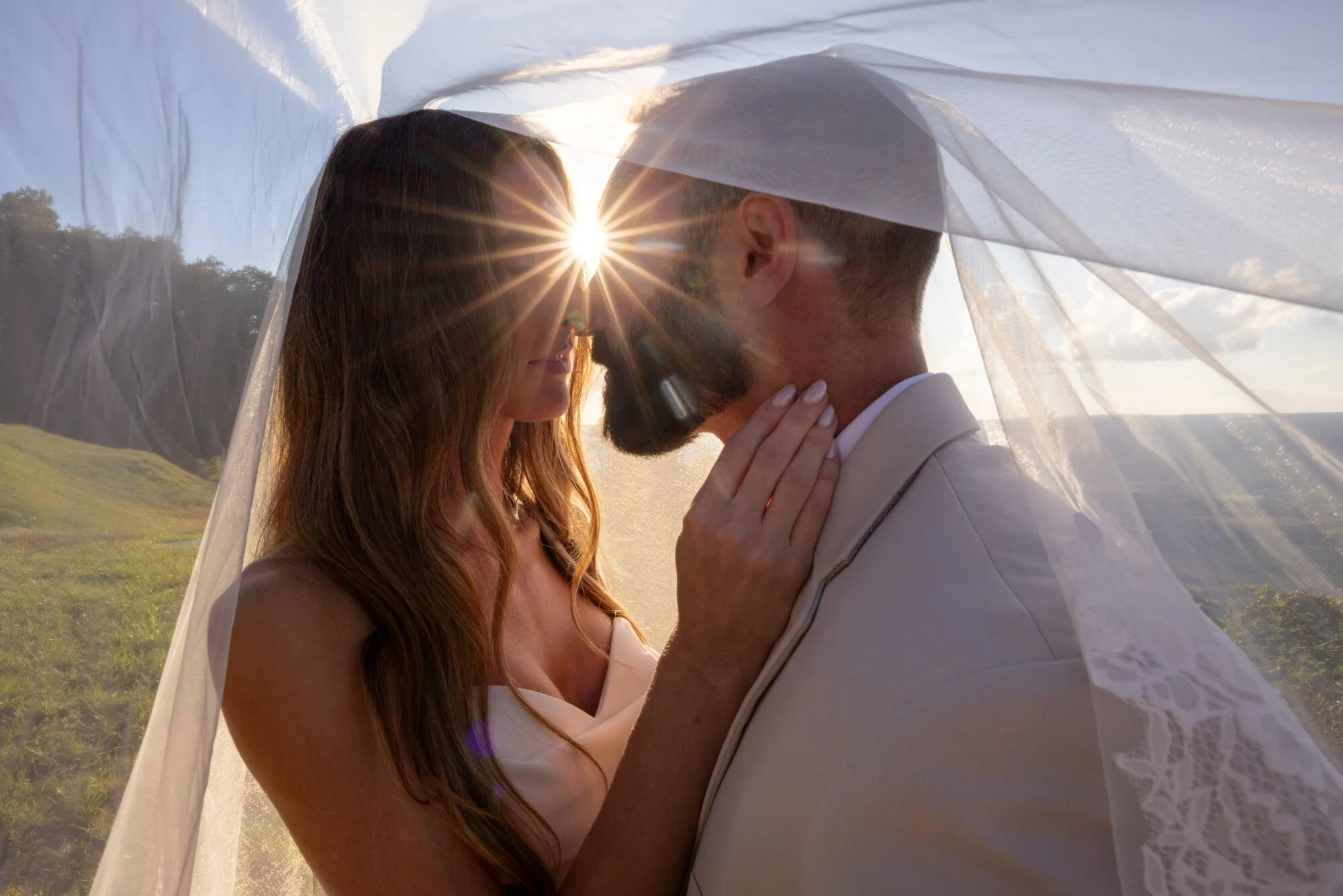 A couple stands close together outdoors under a sheer veil with sunlight creating a starburst effect between their faces, as the woman gently touches the man's neck.