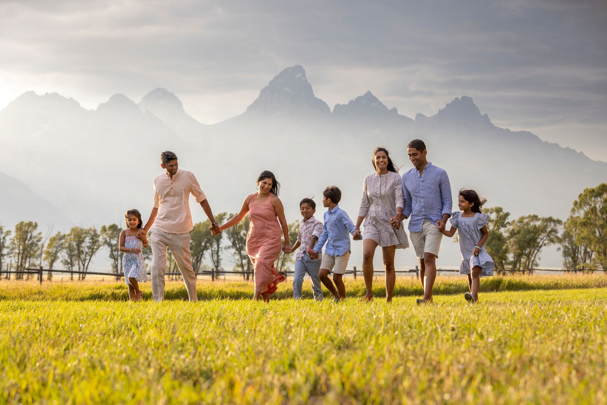A family of nine holding hands and walking through a grassy field, with mountains and cloudy sky in the background.
