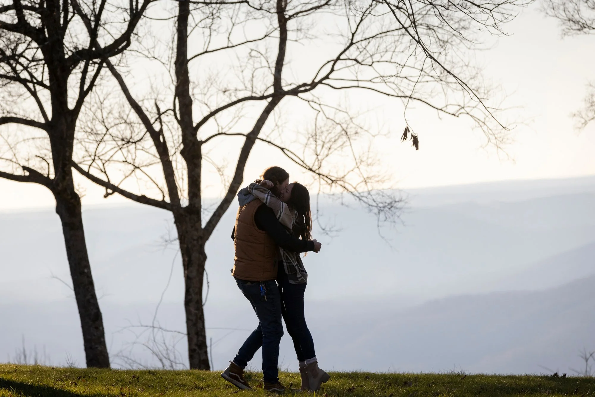 A couple sharing a kiss under the trees near a body of water during sunset or sunrise.