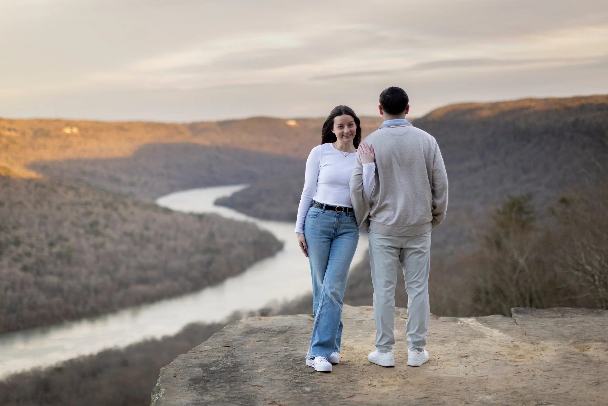 A young woman and a young man walk together along a cliff overlooking a winding river, with hills and a cloudy sky in the background.