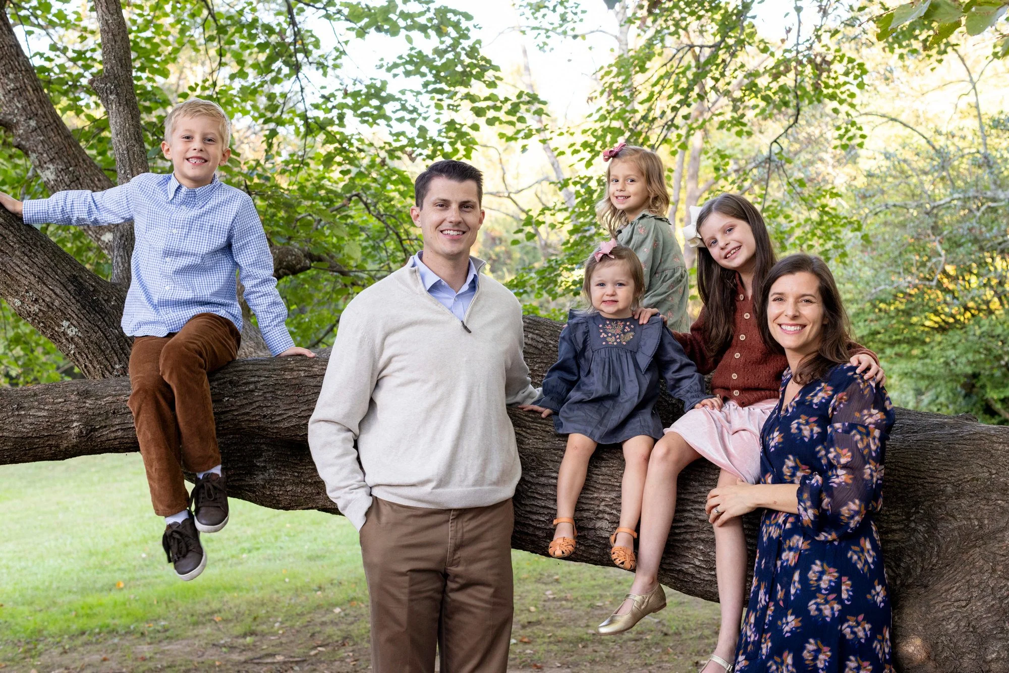 A family of six posing on a large tree branch in a park with green foliage.