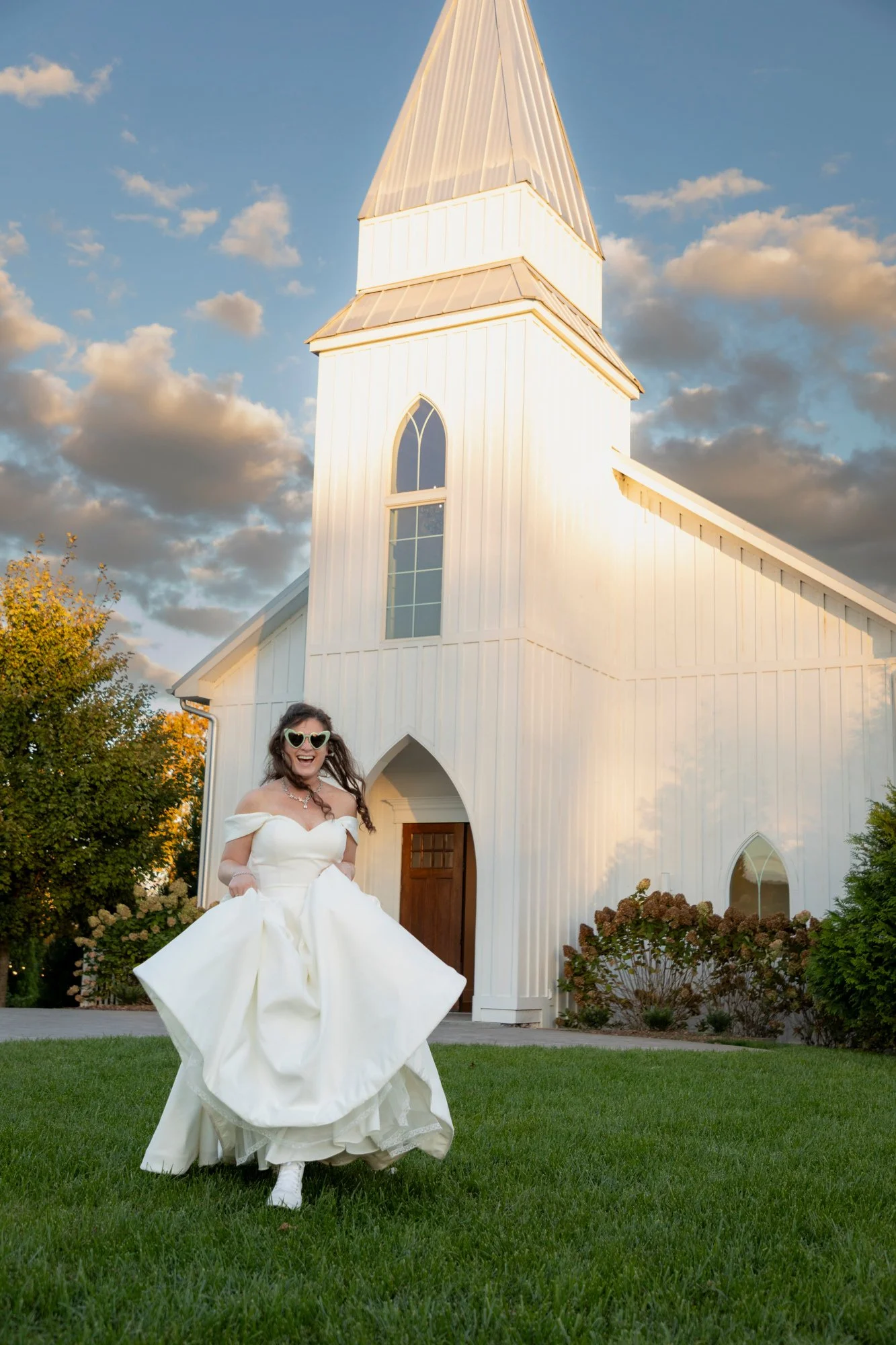 A woman in a white wedding dress holds the skirt of her dress while standing on a green lawn in front of a white church with a tall steeple and arched windows. She is smiling and wearing sunglasses, and the sky behind has scattered clouds at sunset.