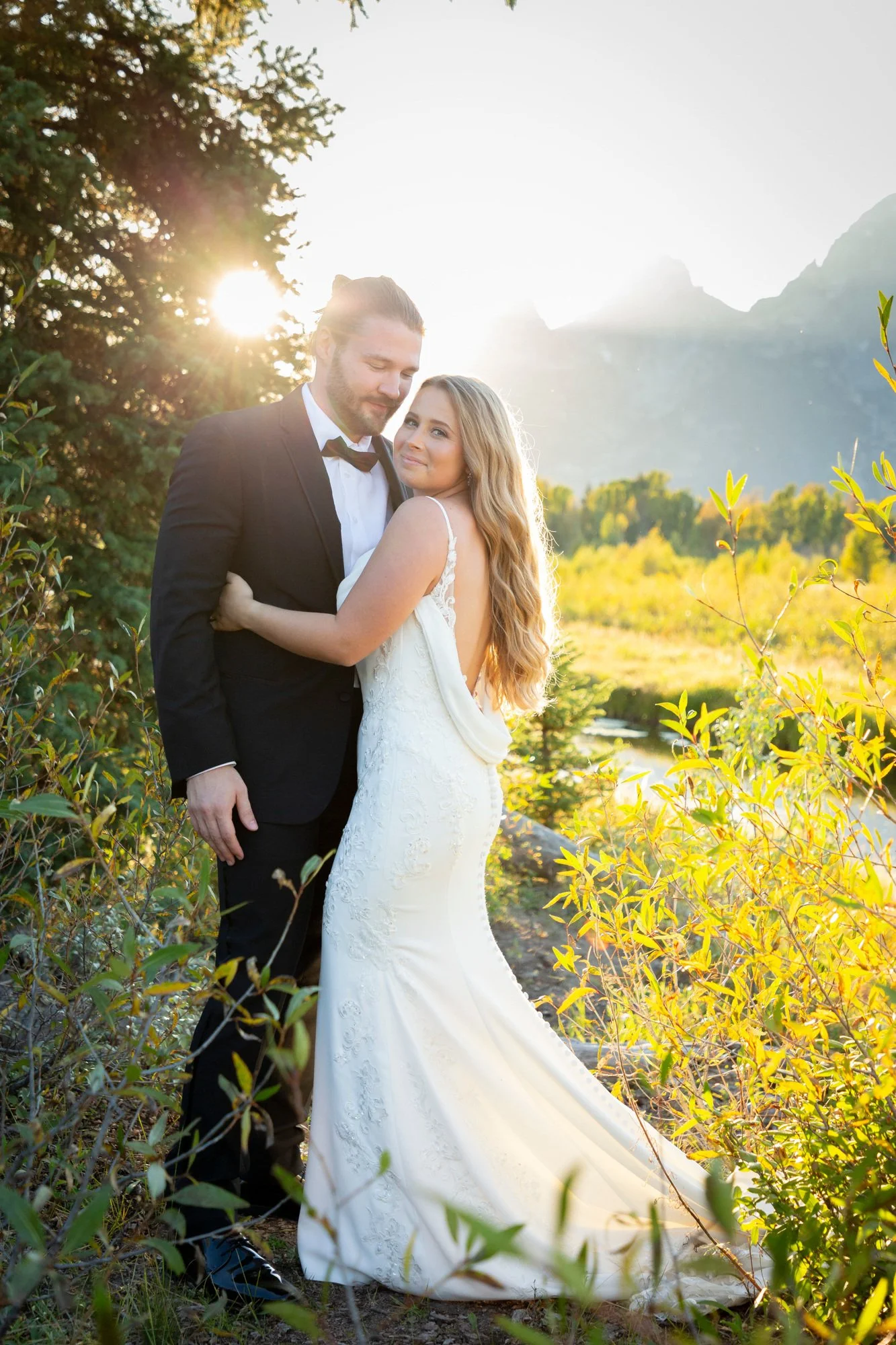 A newlywed couple dressed in wedding attire standing outdoors during sunset in a natural setting with mountains and trees in the background, surrounded by greenery.