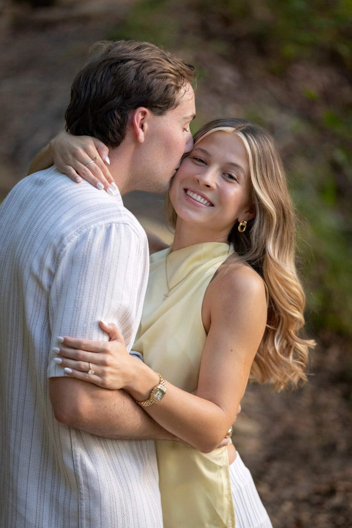 A man is kissing a woman on the cheek while embracing her, both smiling. The woman has long, wavy hair and is wearing gold hoop earrings, a yellow dress, and jewelry. The man has short hair and is wearing a white, textured shirt. They are outdoors wi