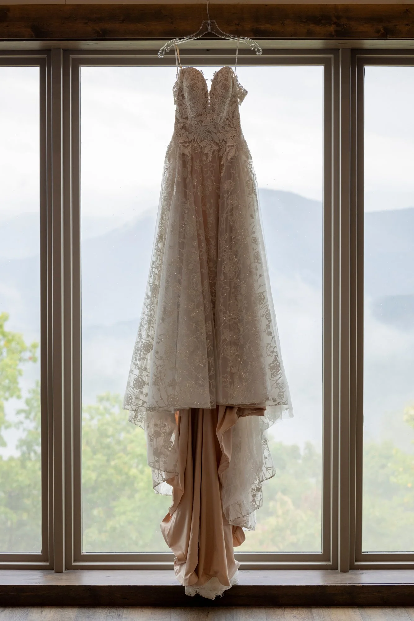 A wedding dress hanging in front of a large window with mountains and trees visible outside.