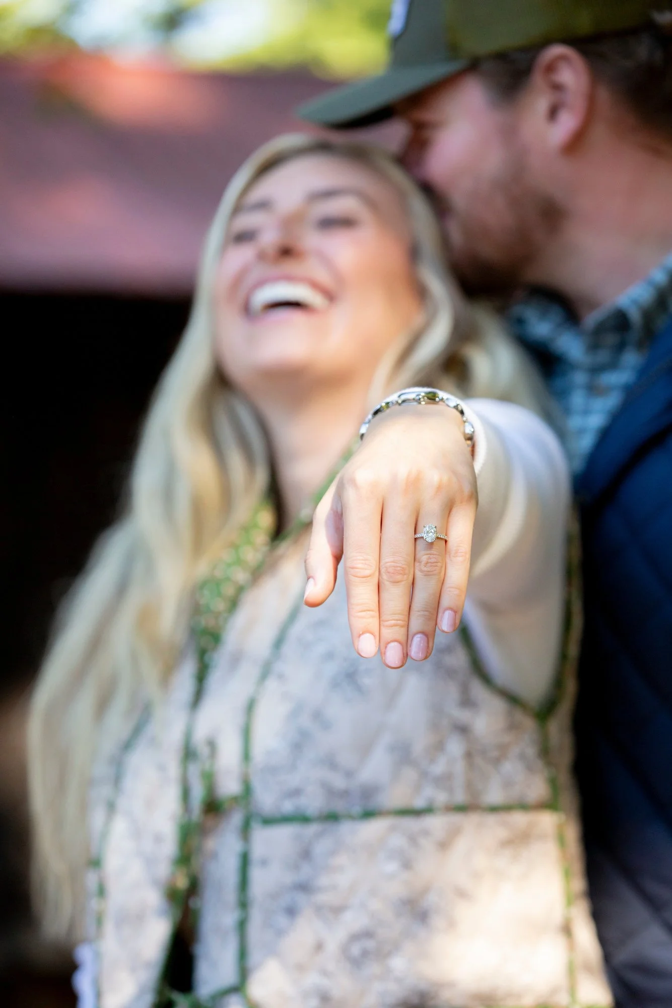 A woman showing her engagement ring with a man kissing her forehead in the background.