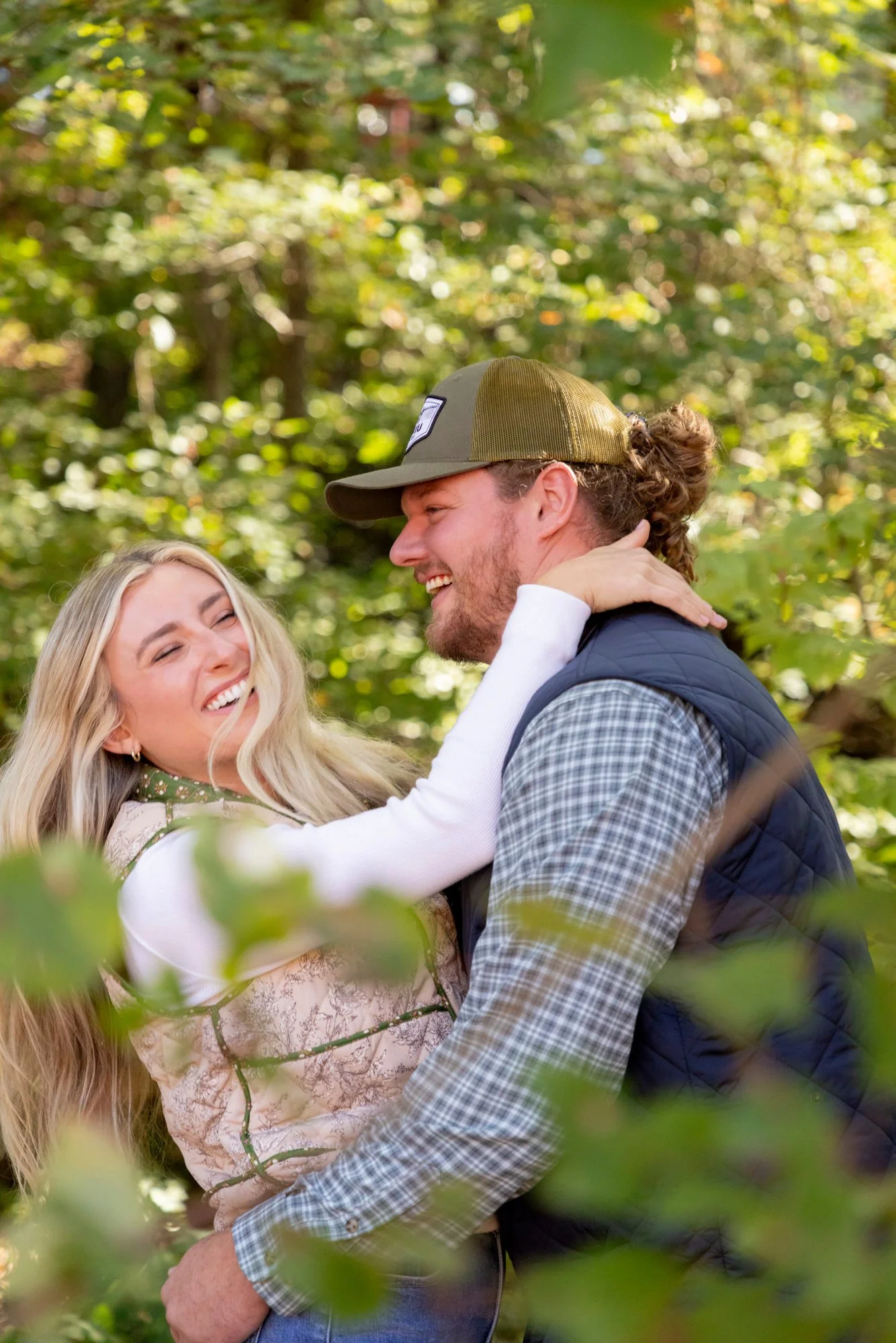 A couple, a woman with long blonde hair and a man with curly hair, are smiling and embracing each other outdoors in a green, leafy environment.