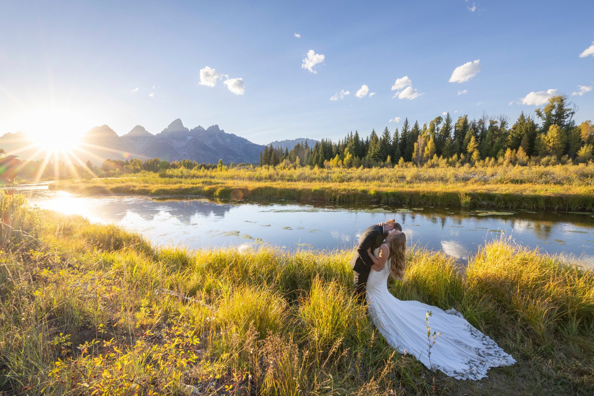 A couple in wedding attire kissing by a river with trees, mountains, and a setting sun in the background.
