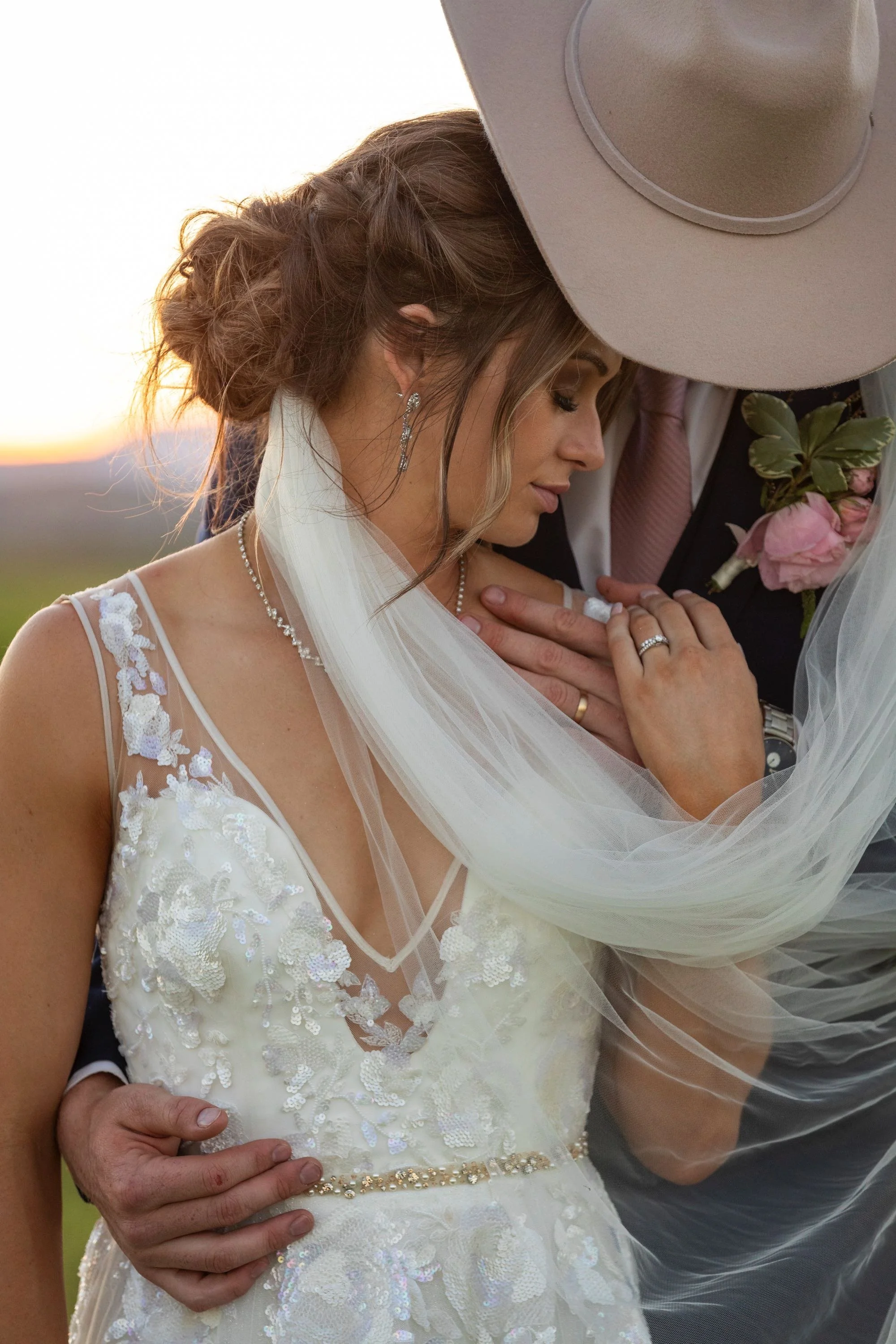 A bride and groom are close together, with the bride's eyes closed and her head leaning against the groom. The bride is wearing a white wedding dress with lace and floral details, earrings, and a necklace. The groom is wearing a dark suit with a ligh