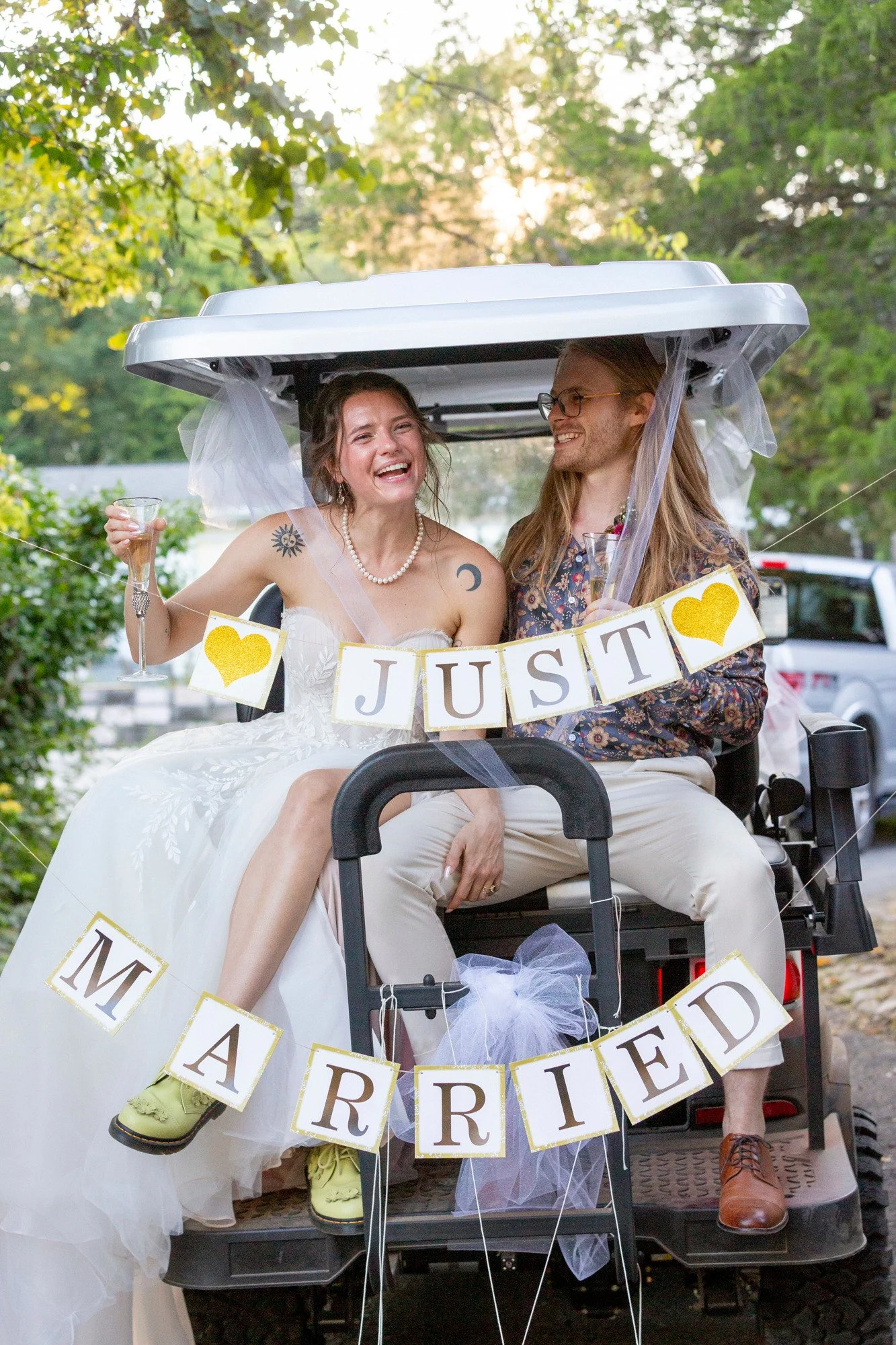 A newlywed couple sits on a golf cart decorated with a banner that says 'JUST MARRIED' and cheering red hearts. The bride is wearing a white wedding dress and pearl necklace, holding a glass of champagne with a big smile. The groom wears glasses and 