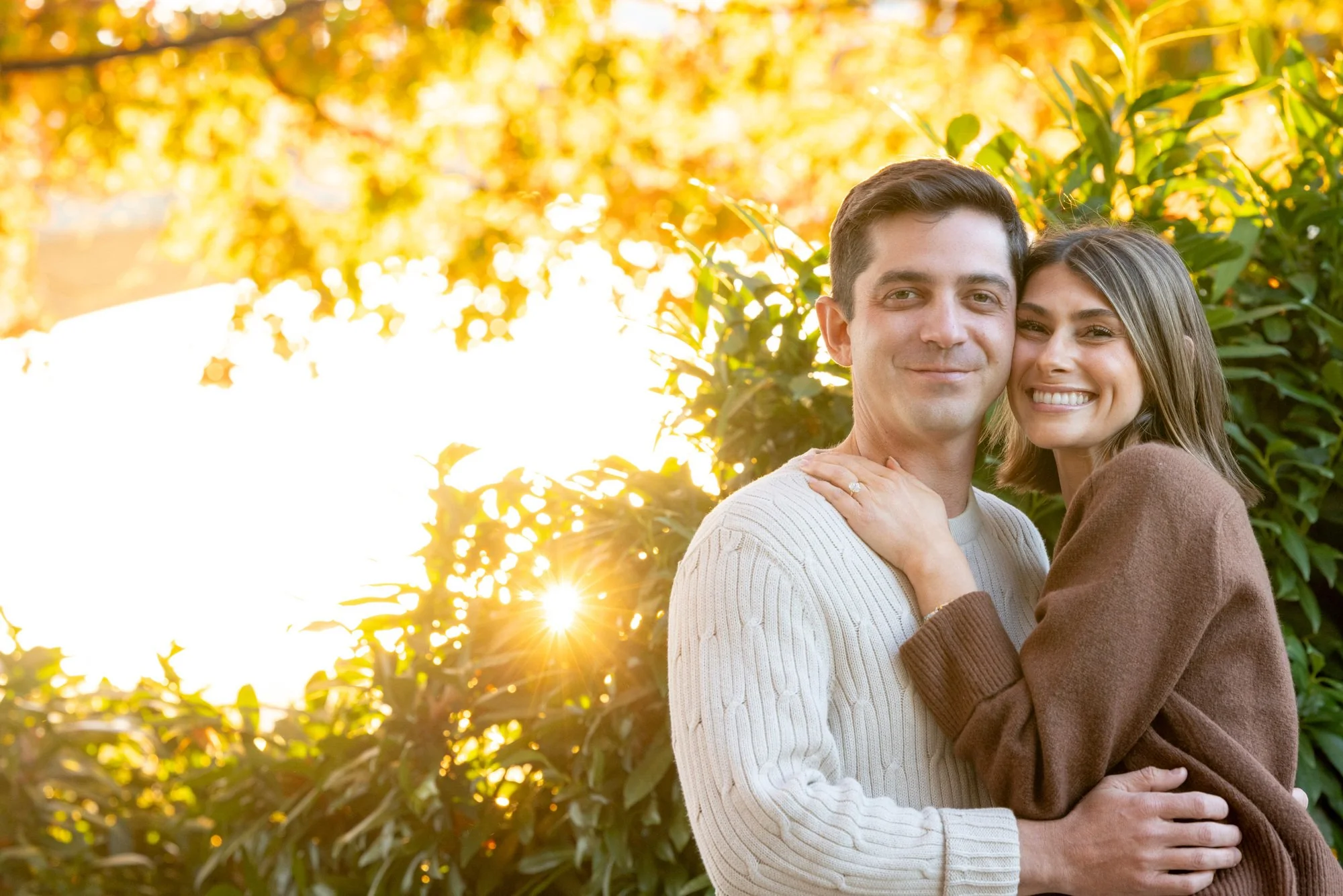 A happy couple embracing outdoors during sunset, with trees and sunlight in the background.