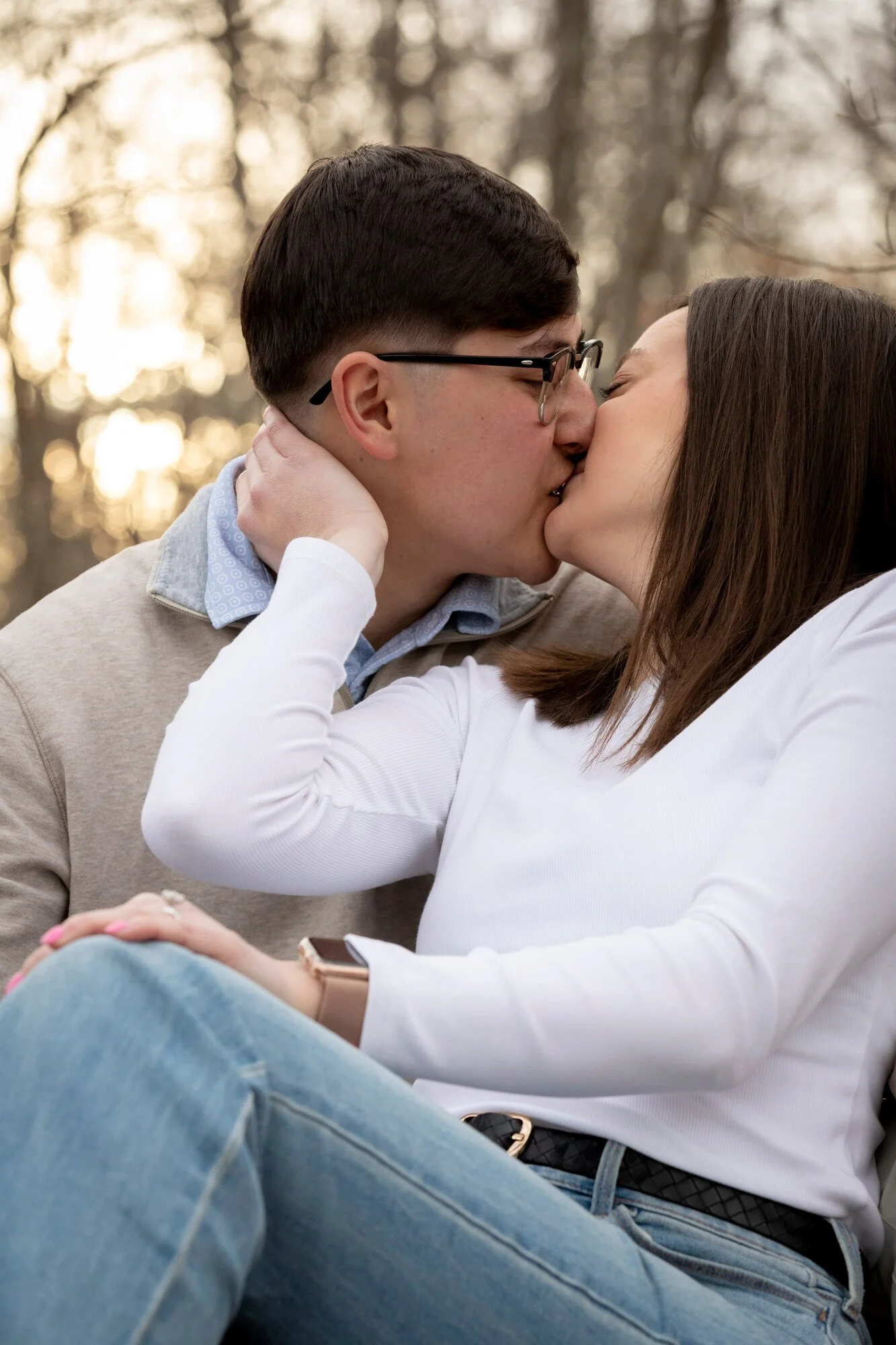 A couple kissing outdoors, with the person in front wearing blue jeans and a white shirt. The person in the background has short dark hair, glasses, and is wearing a light-colored sweater. The background shows trees with blurred light.