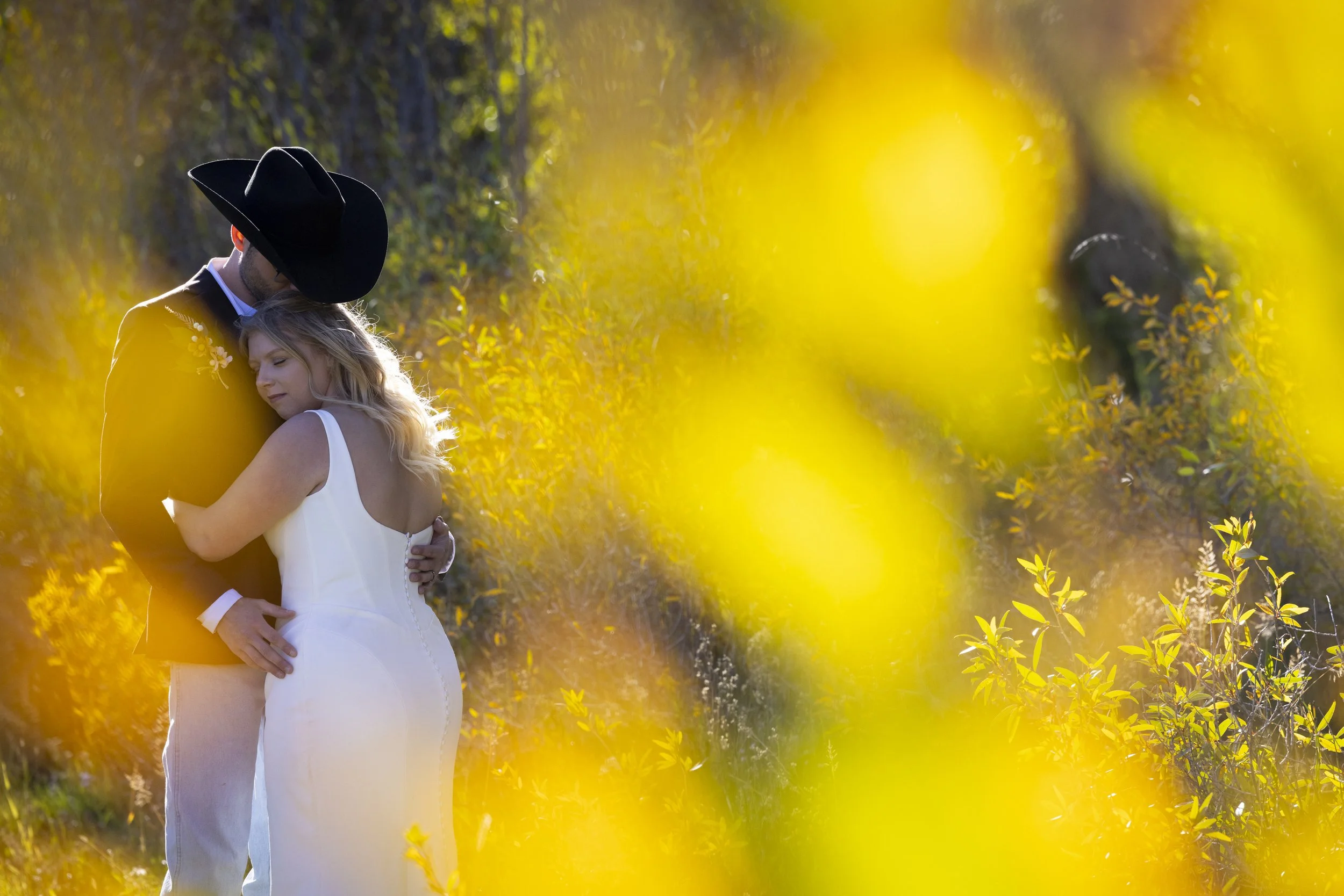 A couple dressed in wedding attire embracing outdoors during sunset, surrounded by yellow flowers and greenery.