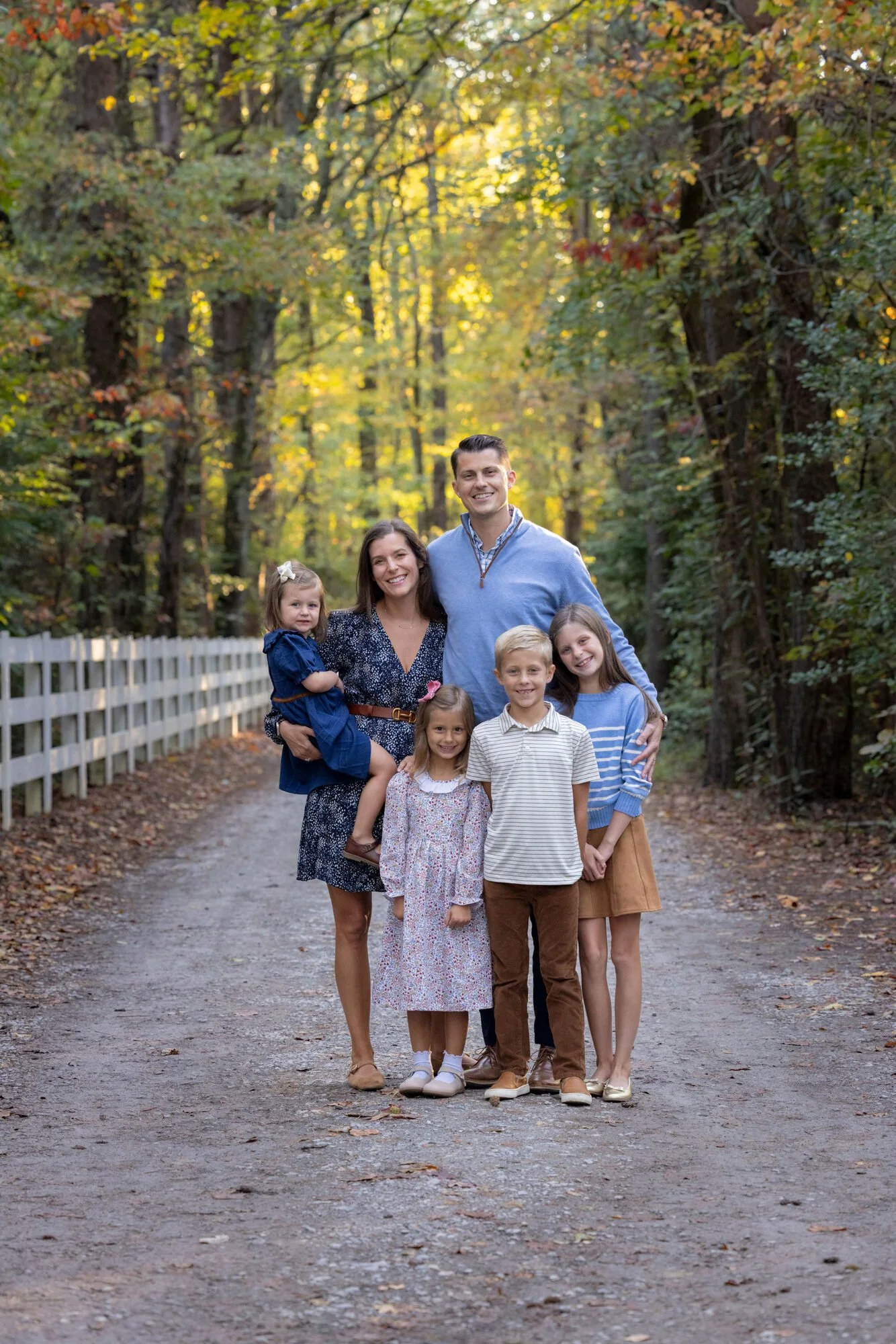 A family of seven standing on a dirt path in a wooded area with autumn foliage. The family includes two adults and five children, all smiling at the camera.